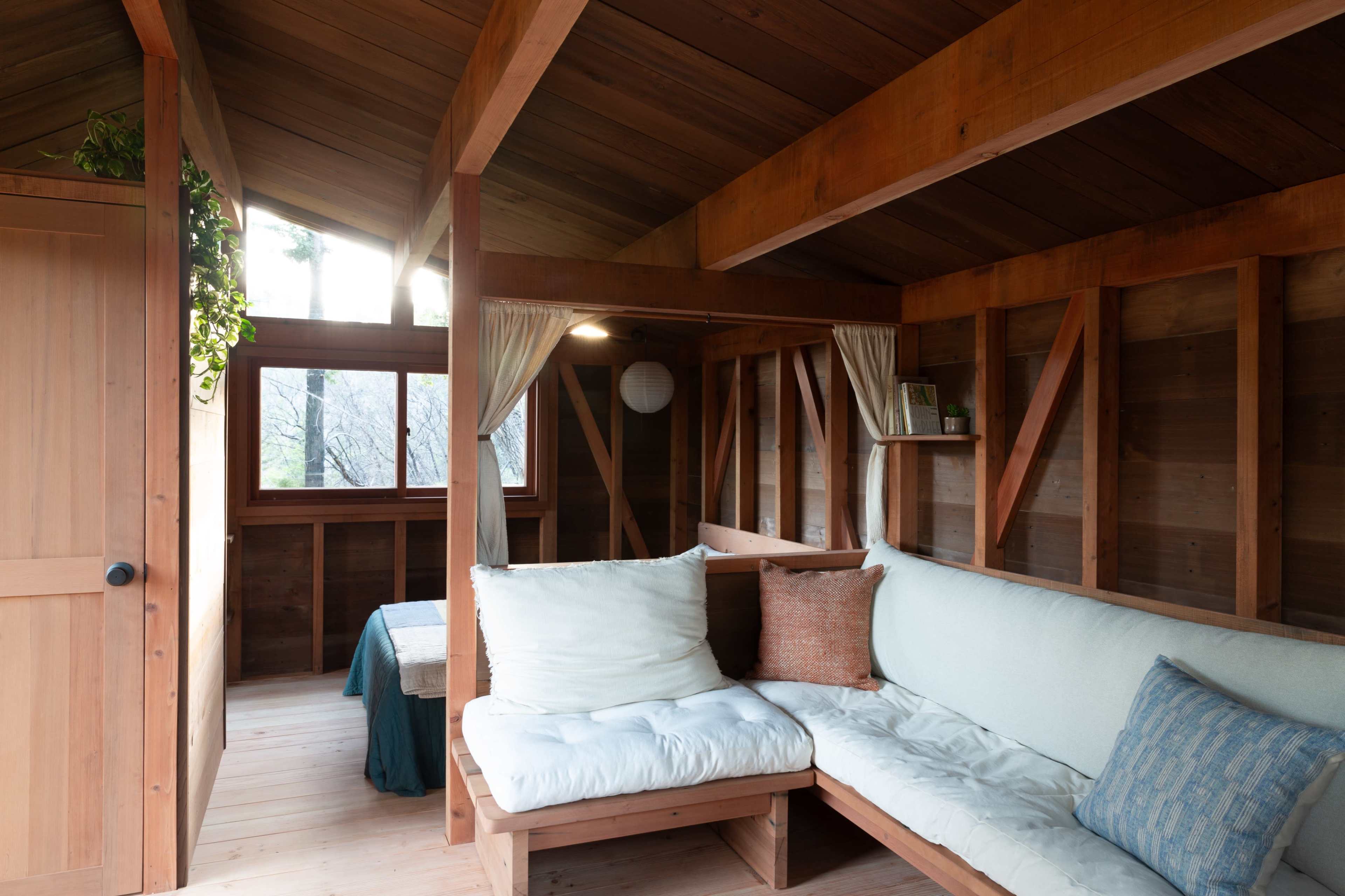 A wooden interior of a small cabin with a couch, a table, and large windows allowing natural light to enter.