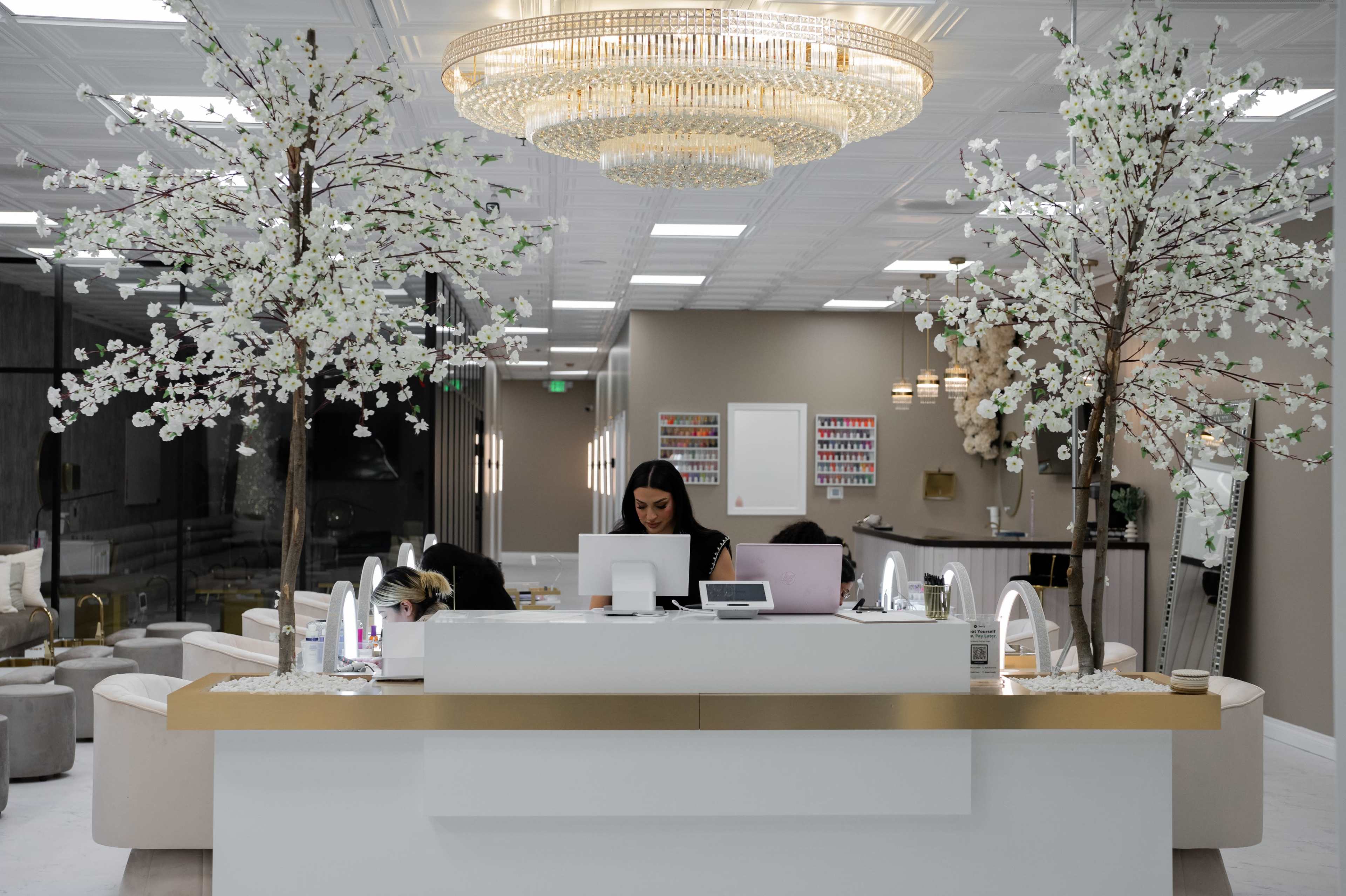 A modern salon reception area features a white front desk adorned with decorative cherry blossom trees and a large chandelier overhead.