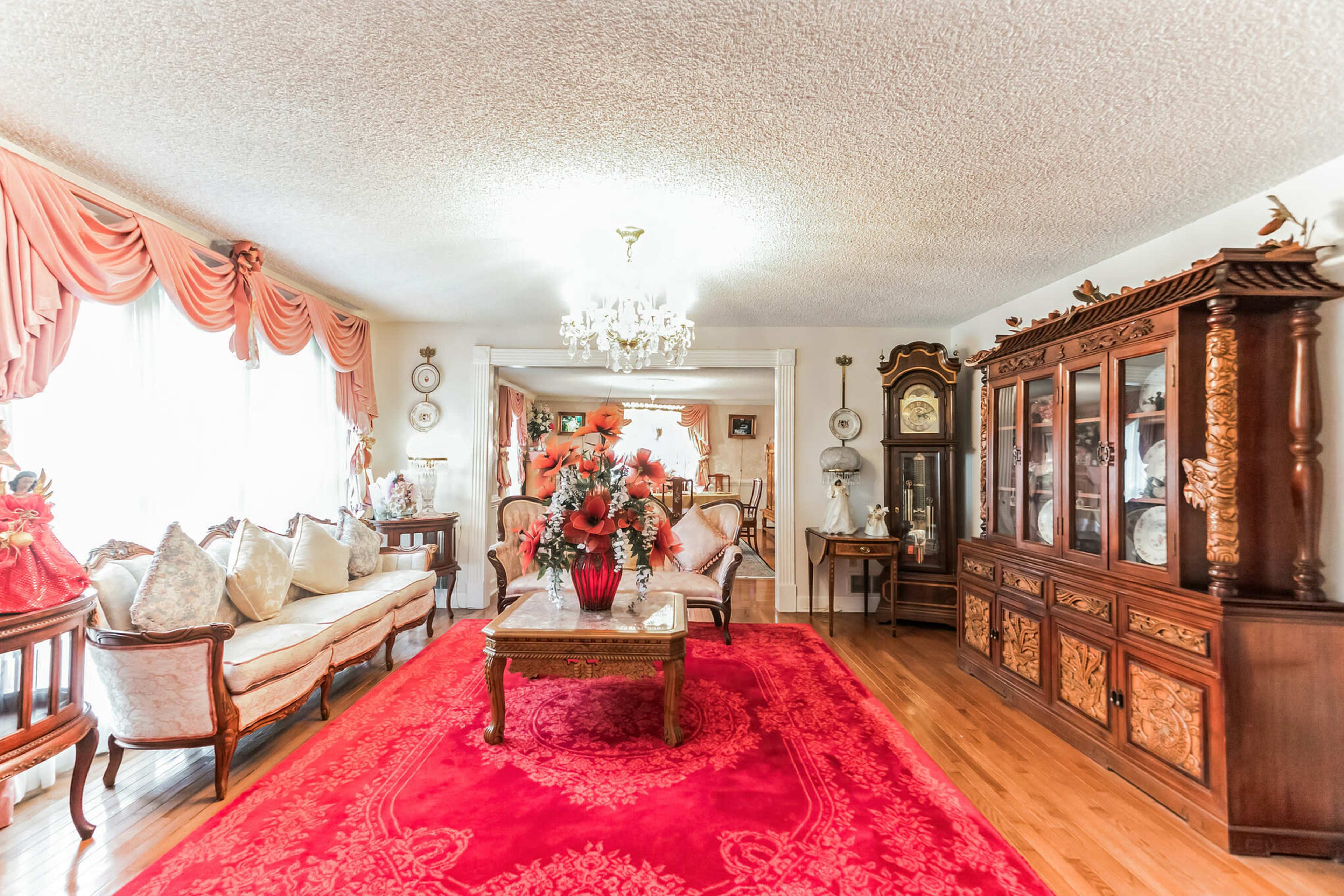 A living room with a large red area rug, a wooden coffee table adorned with flowers, and elegant furniture arranged around a chandelier.