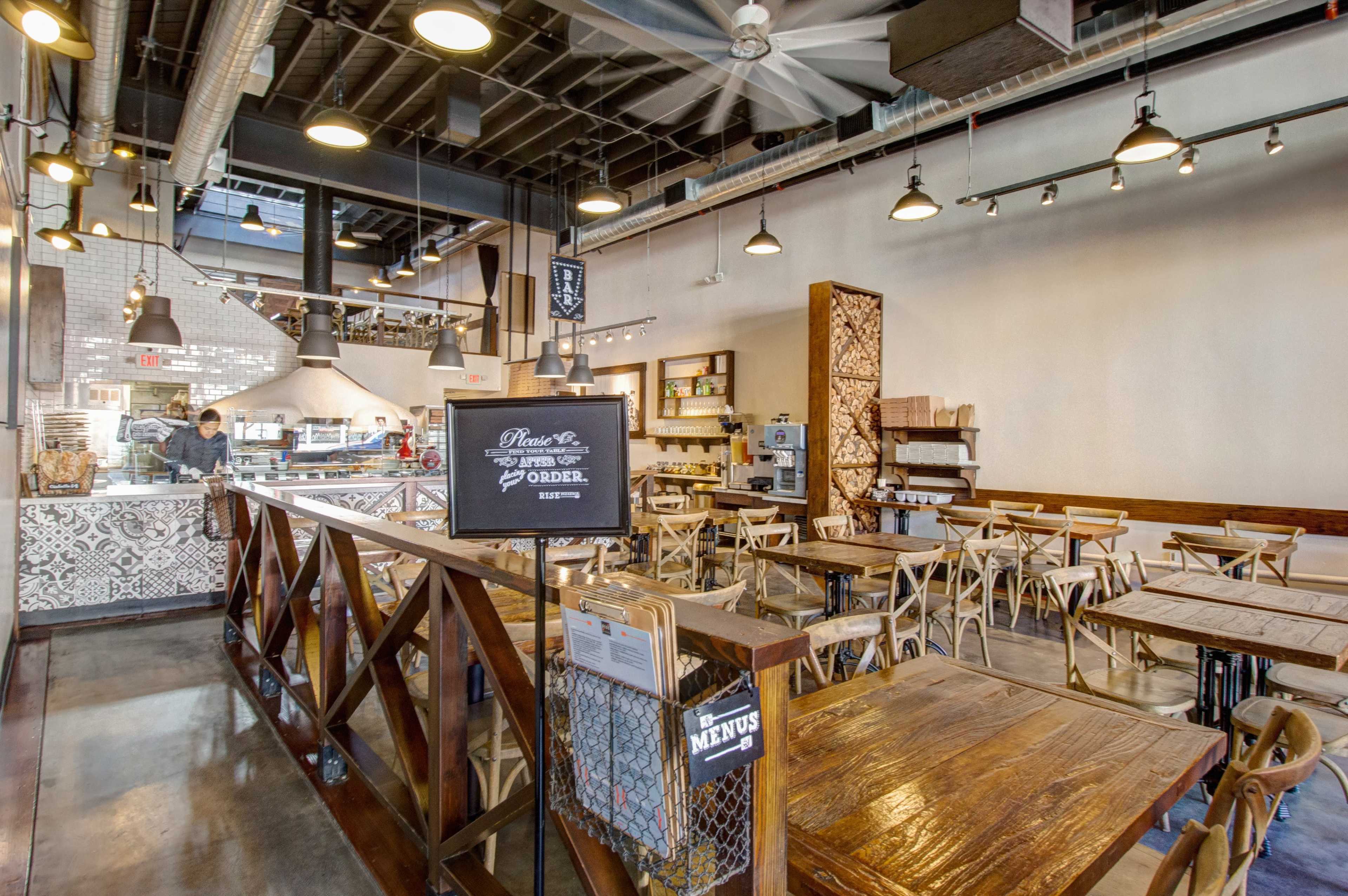 A spacious restaurant interior featuring wooden tables and chairs, a decorative counter, and industrial-style lighting.