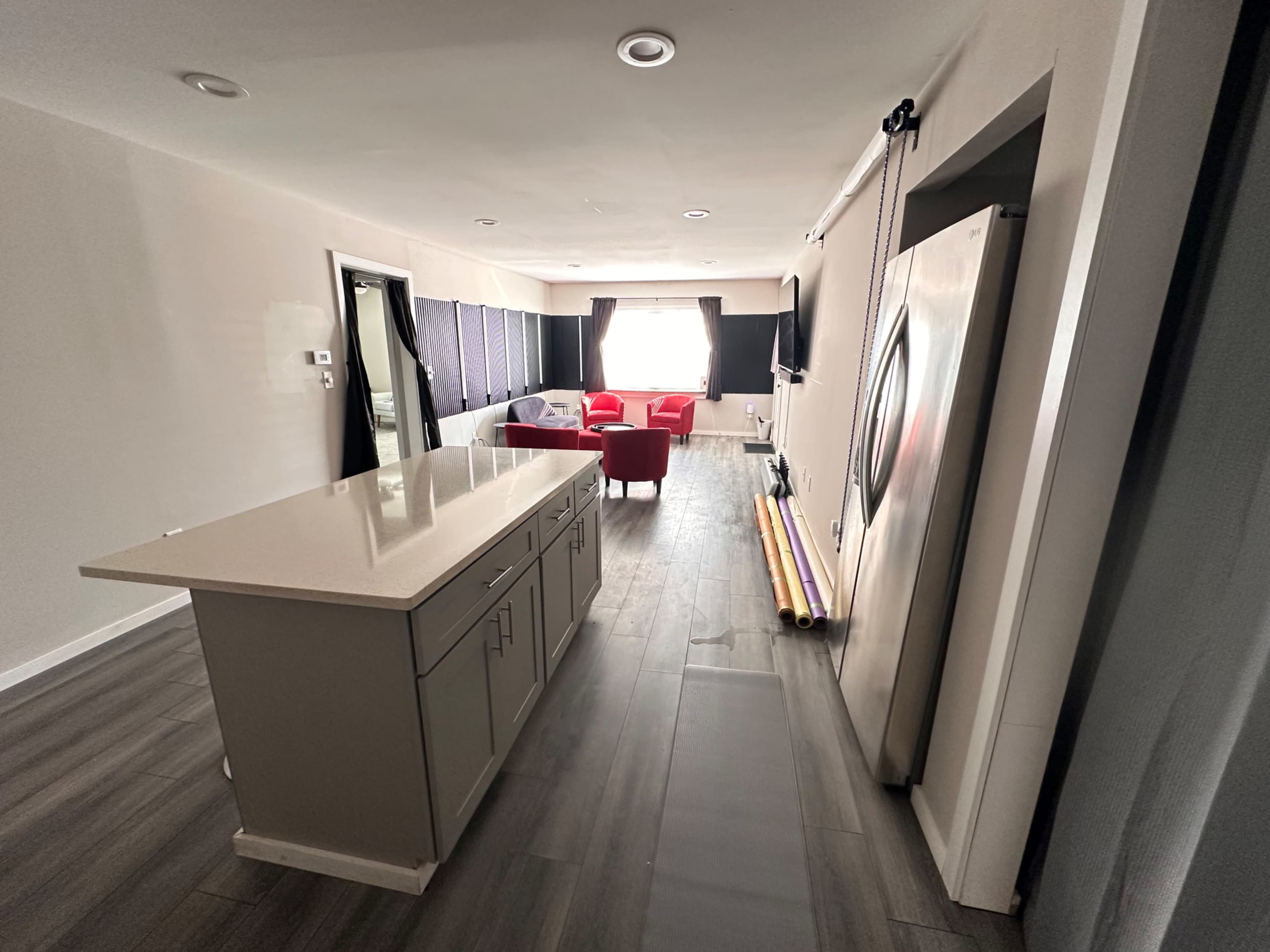 The image shows a modern kitchen area with a central island, leading towards a living room featuring two red chairs and large windows covered with dark curtains.