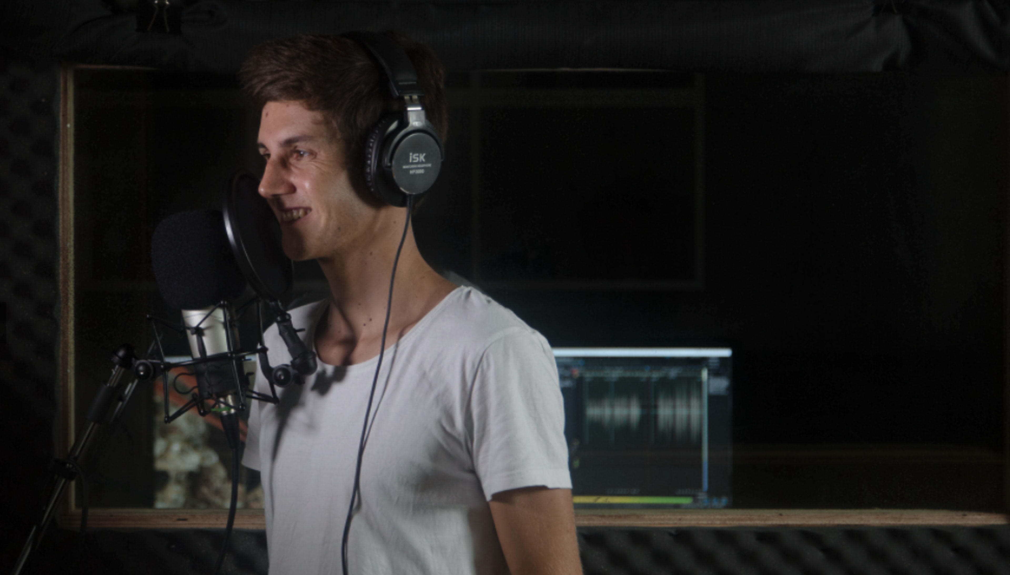 A young man wearing headphones stands in a recording studio, speaking into a microphone.