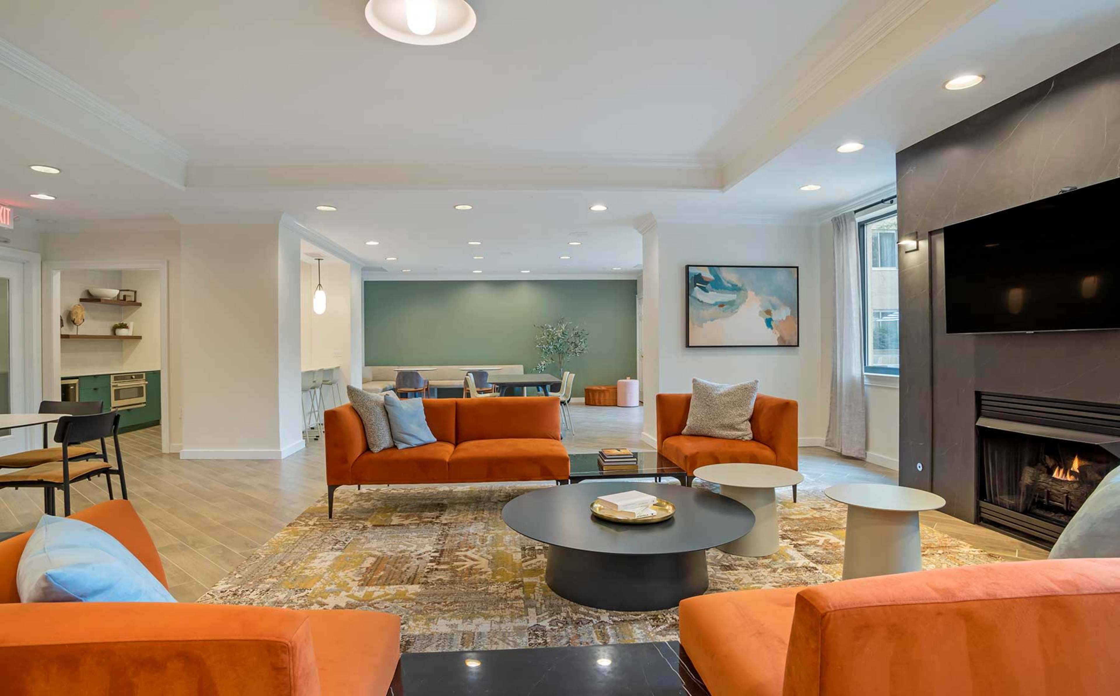 A modern living room features orange couches, a round black coffee table, and a fireplace, with a dining area visible in the background.