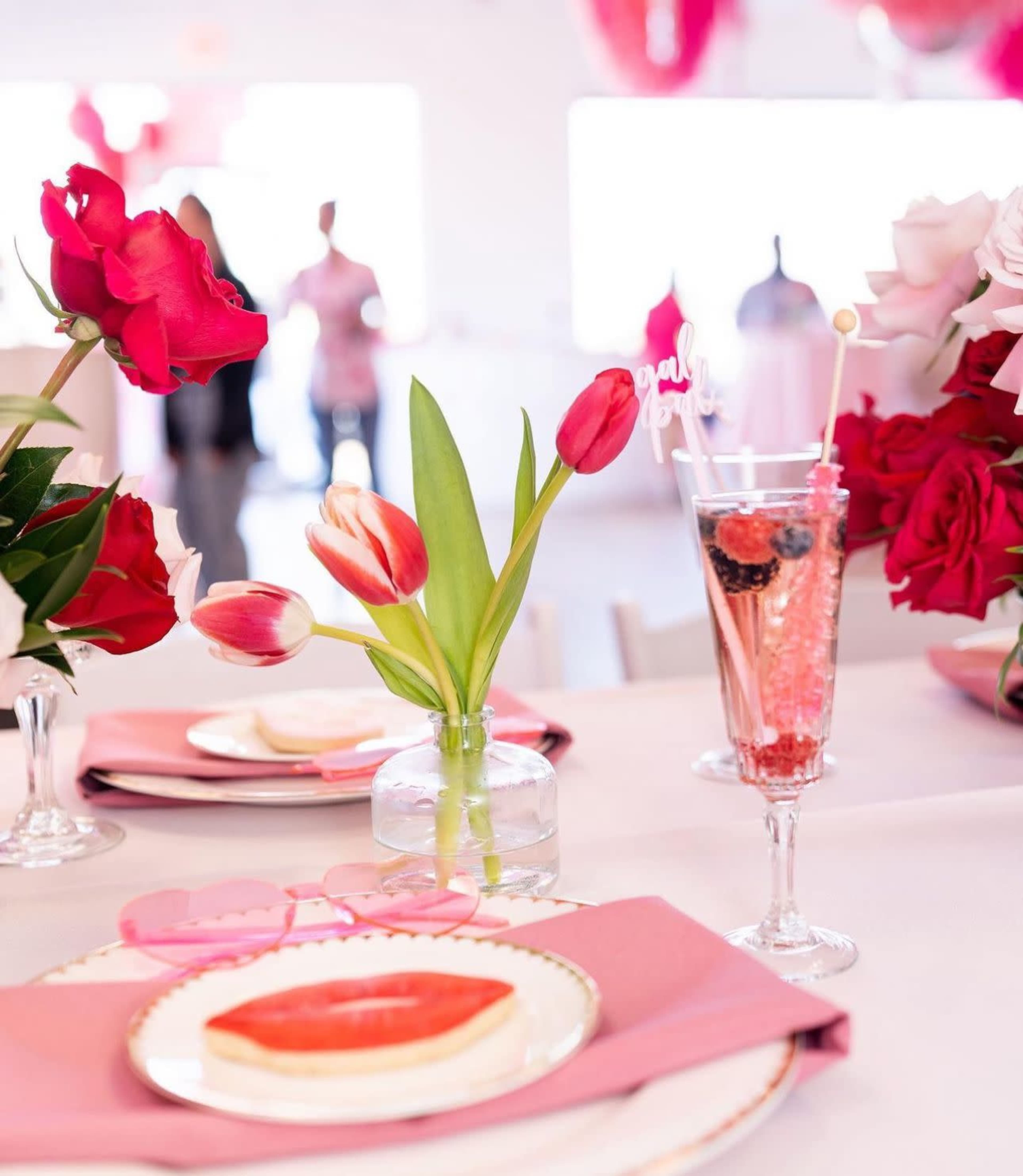 The table is set with pink plates, a glass of pink drink garnished with berries, and a vase of colorful tulips amidst floral decorations.