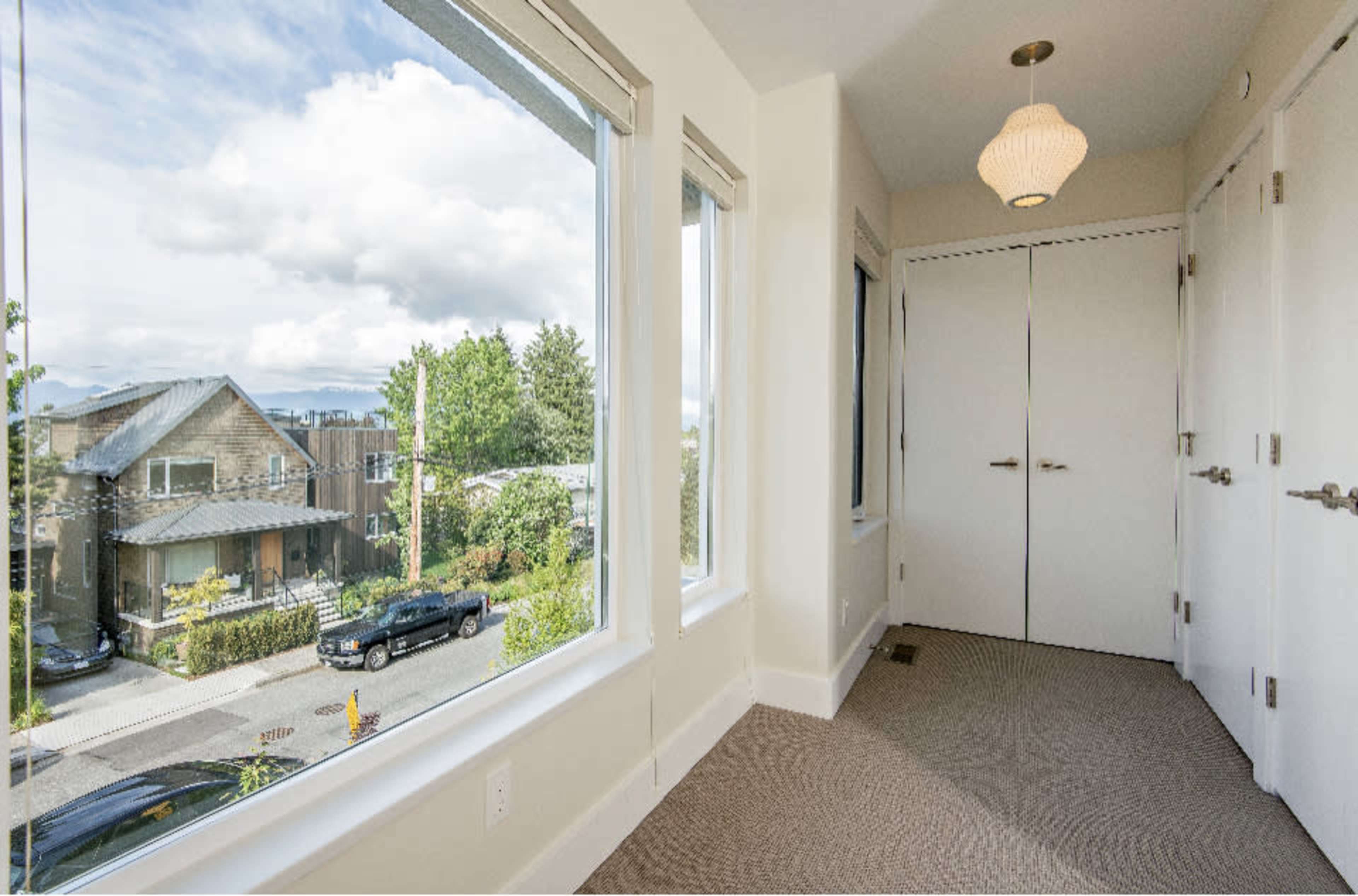 A brightly lit hallway features large windows overlooking a residential area with parked cars and greenery outside.