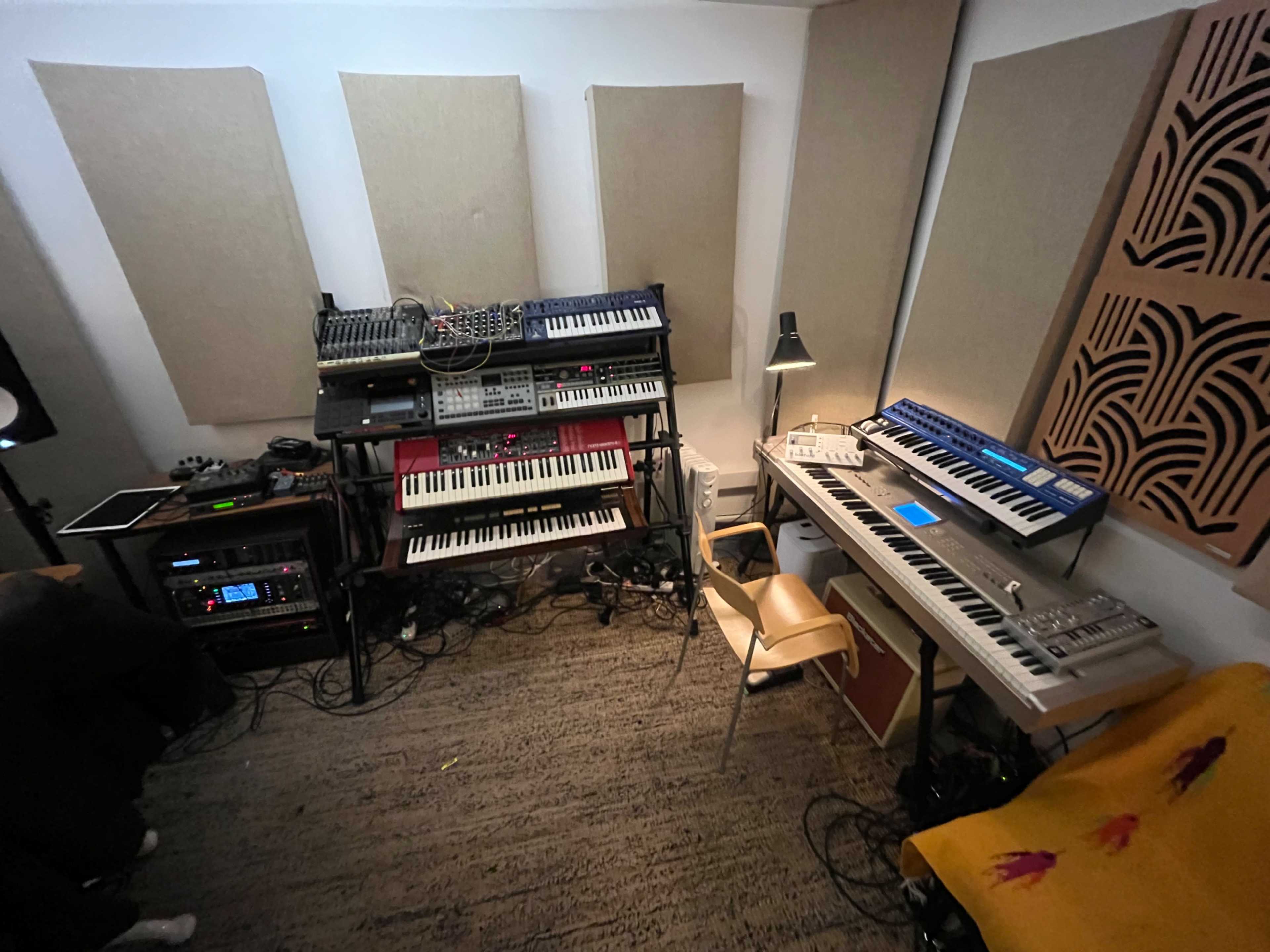 The image shows a home music studio featuring various synthesizers, keyboards, and audio equipment against a backdrop of soundproofing panels.