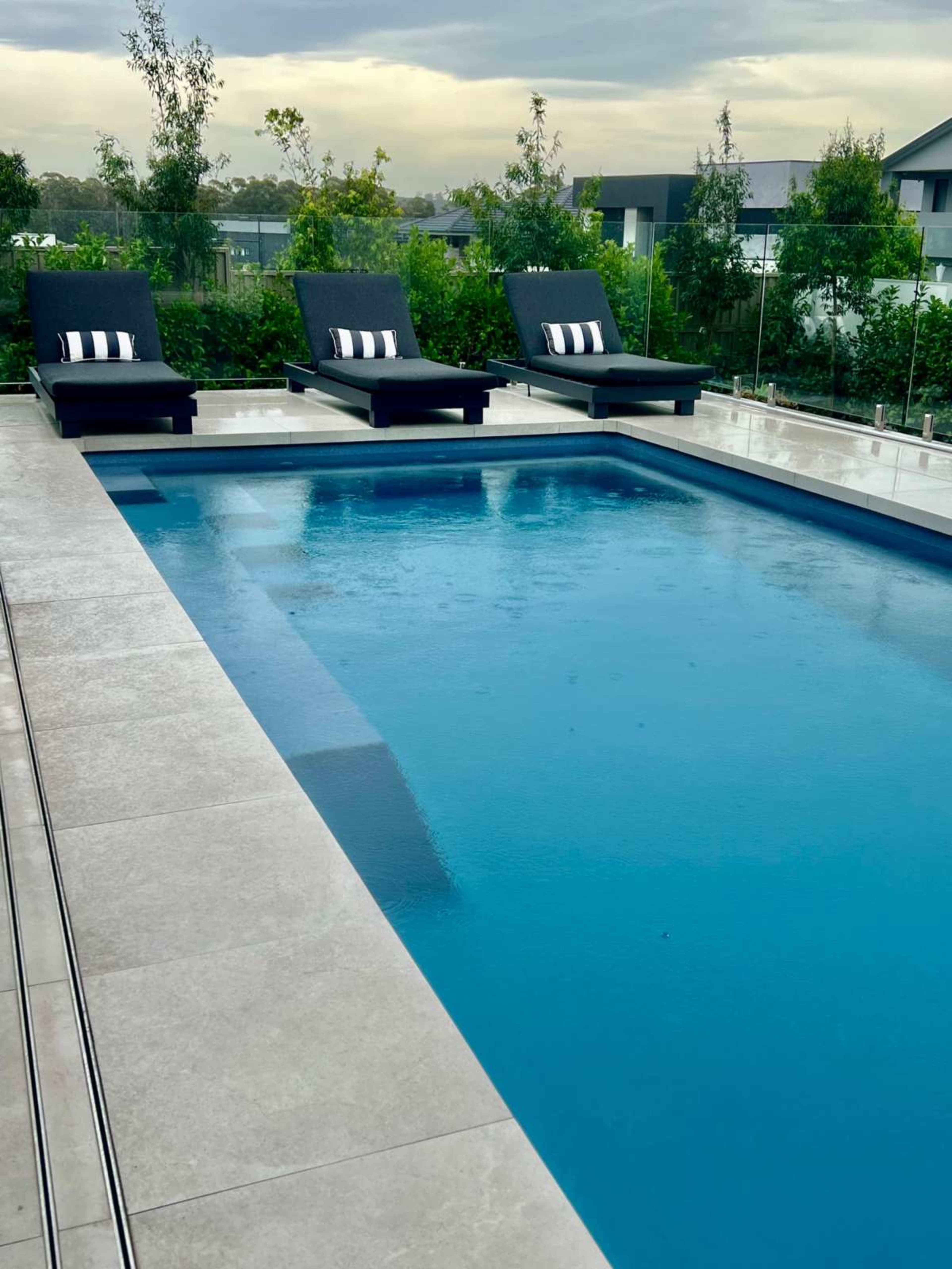 An outdoor swimming pool with three chaise lounges on the deck and greenery in the background.
