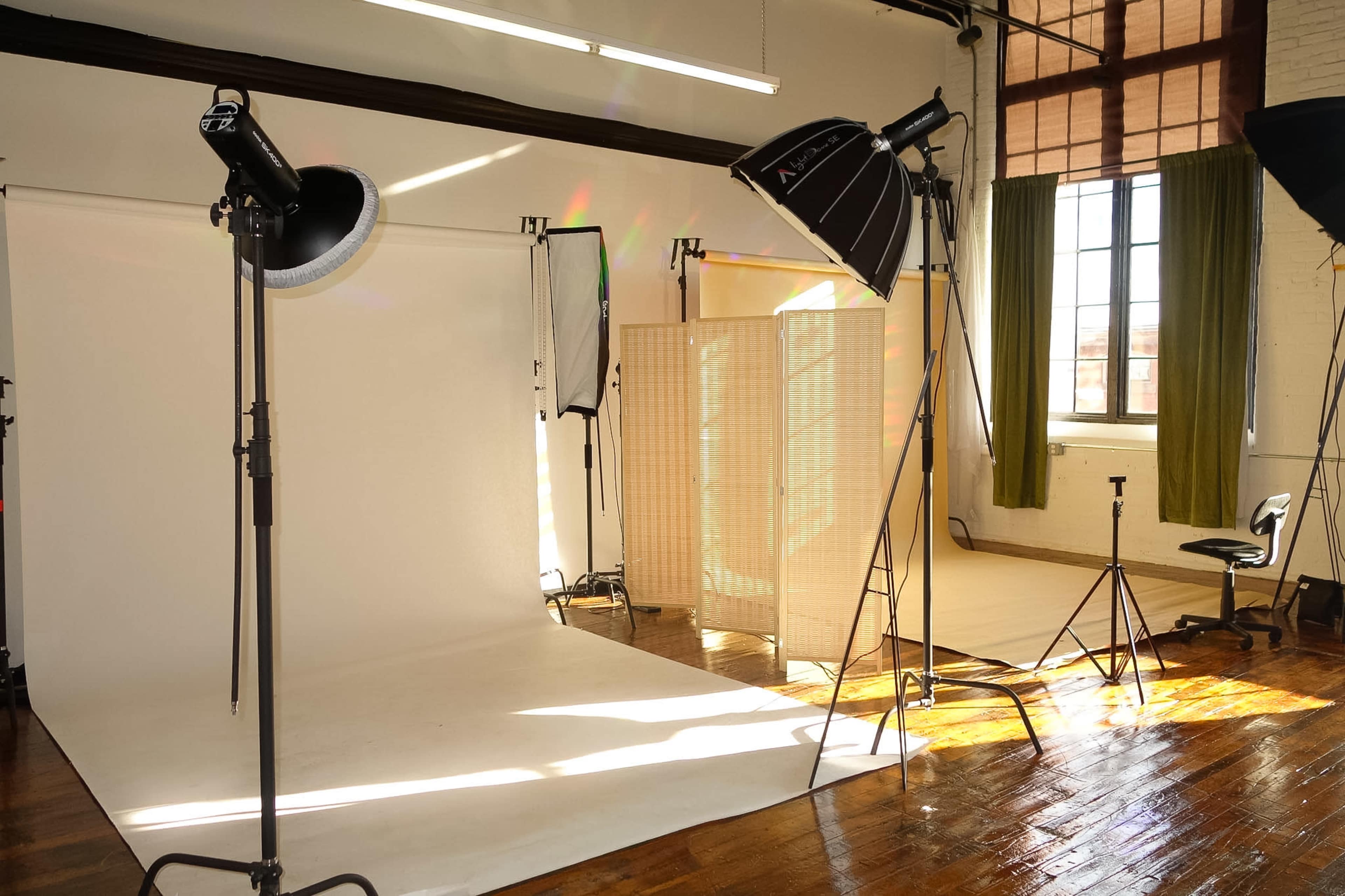 The image shows a photography studio with a white backdrop, light stands, and a folding screen, illuminated by natural light coming through large windows.