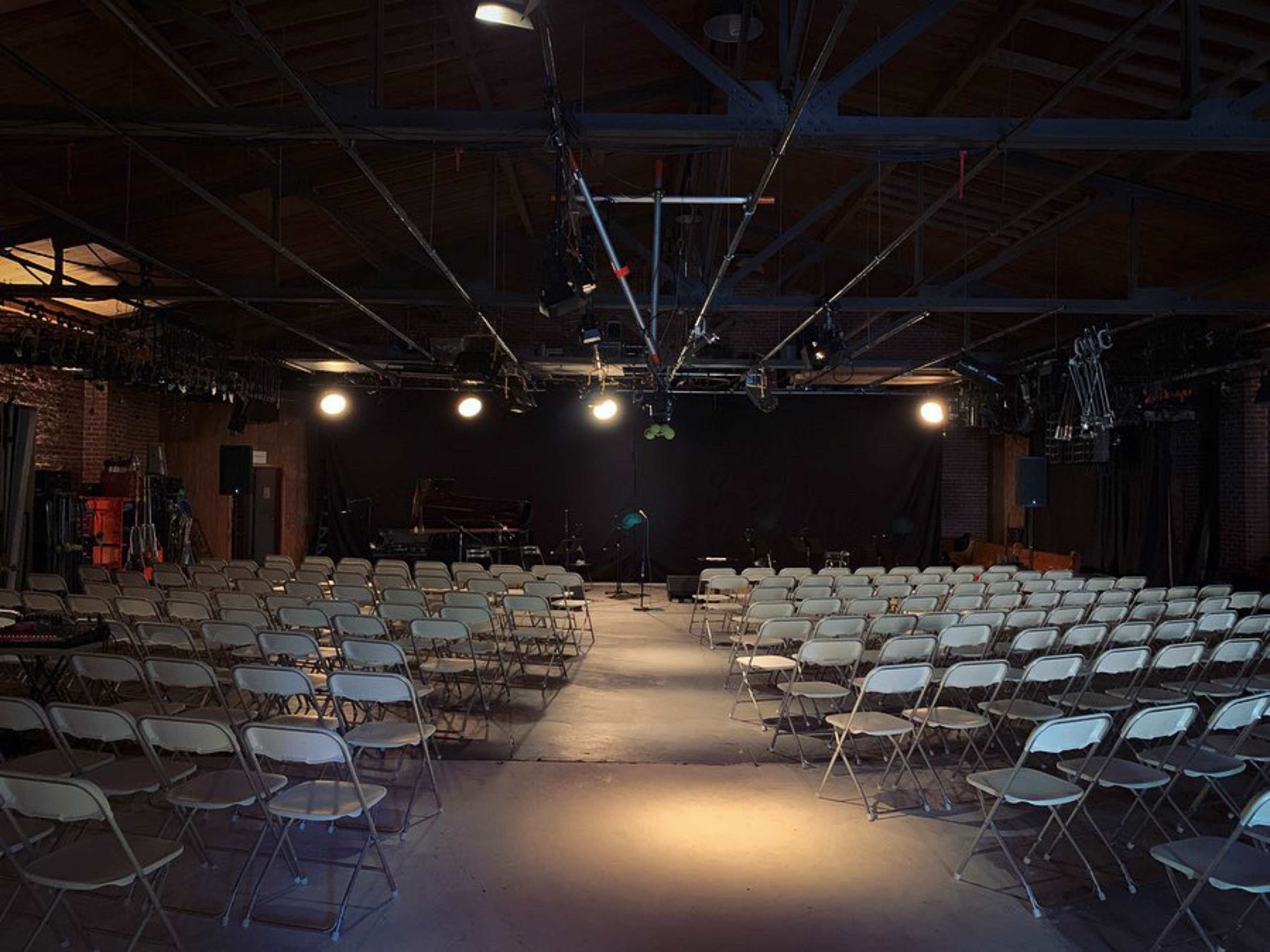 An empty performance venue with rows of white folding chairs facing a small stage equipped with a microphone and other performance equipment.