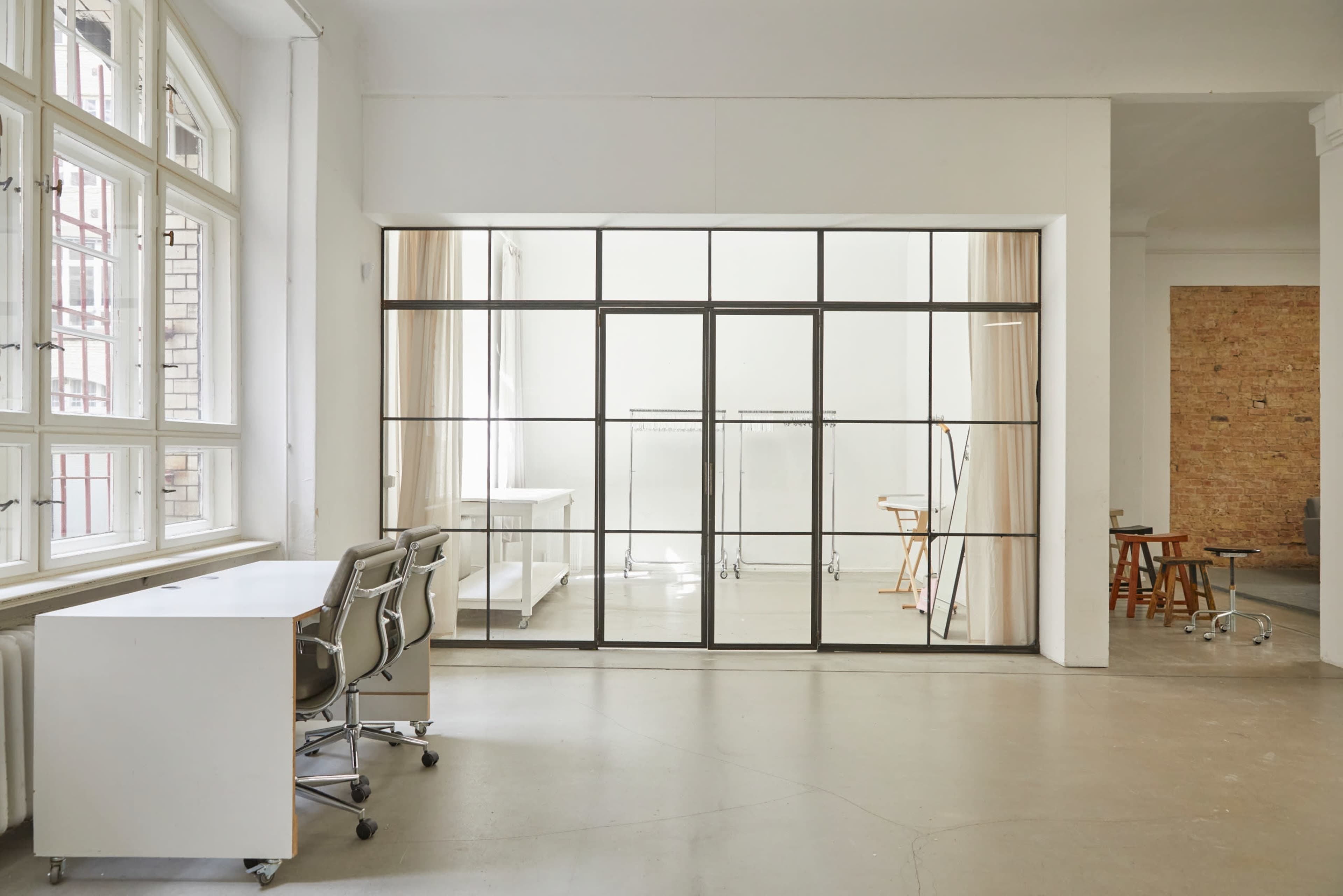 A bright, minimalist office space features large windows, glass paneling, and simple furniture arrangements.