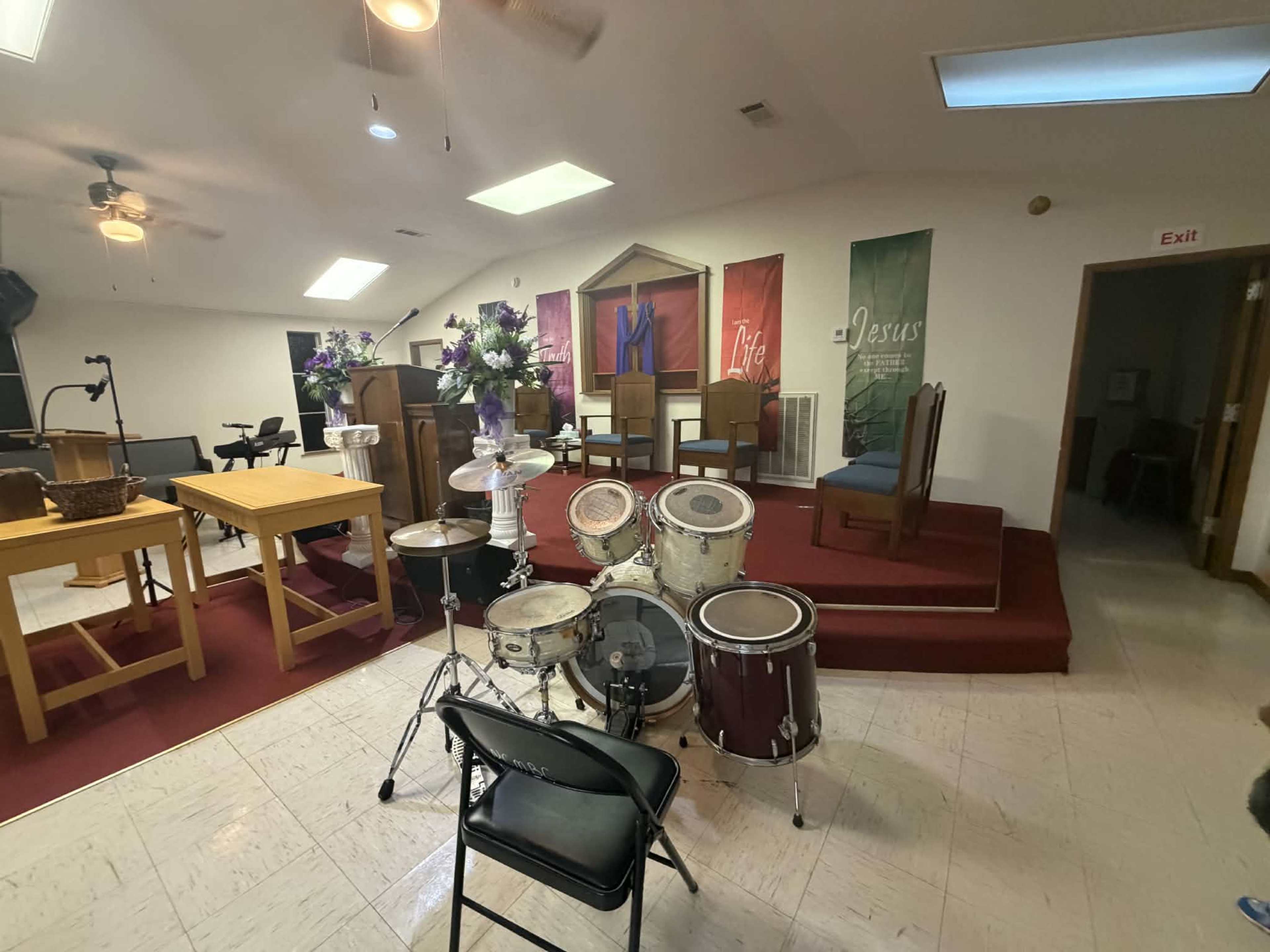 The image shows the interior of a church with a drum set in the foreground and pews arranged in a worship area.