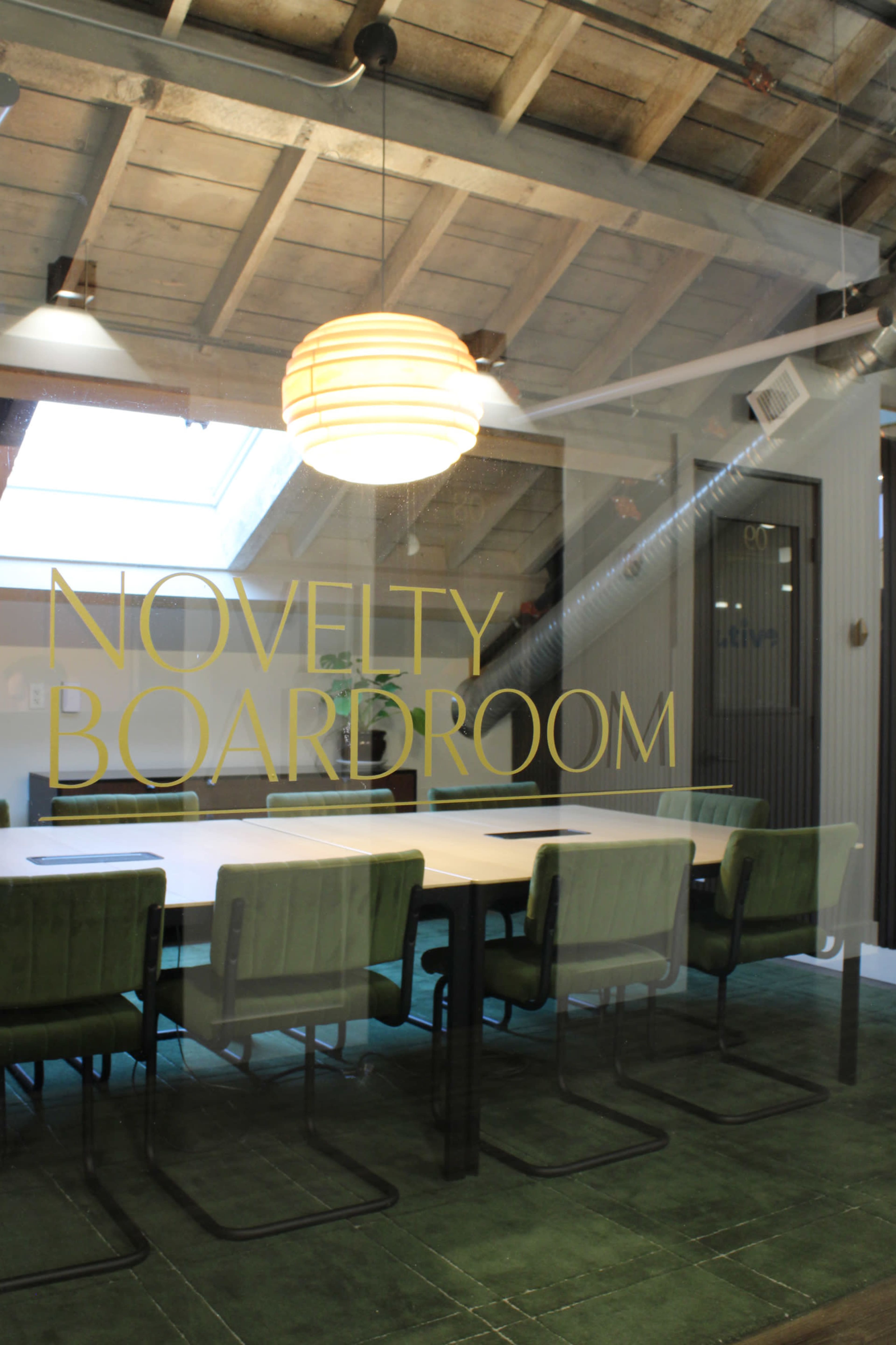 A modern conference room features a large table surrounded by green chairs, with the words "NOVELTY BOARDROOM" displayed on a glass wall.