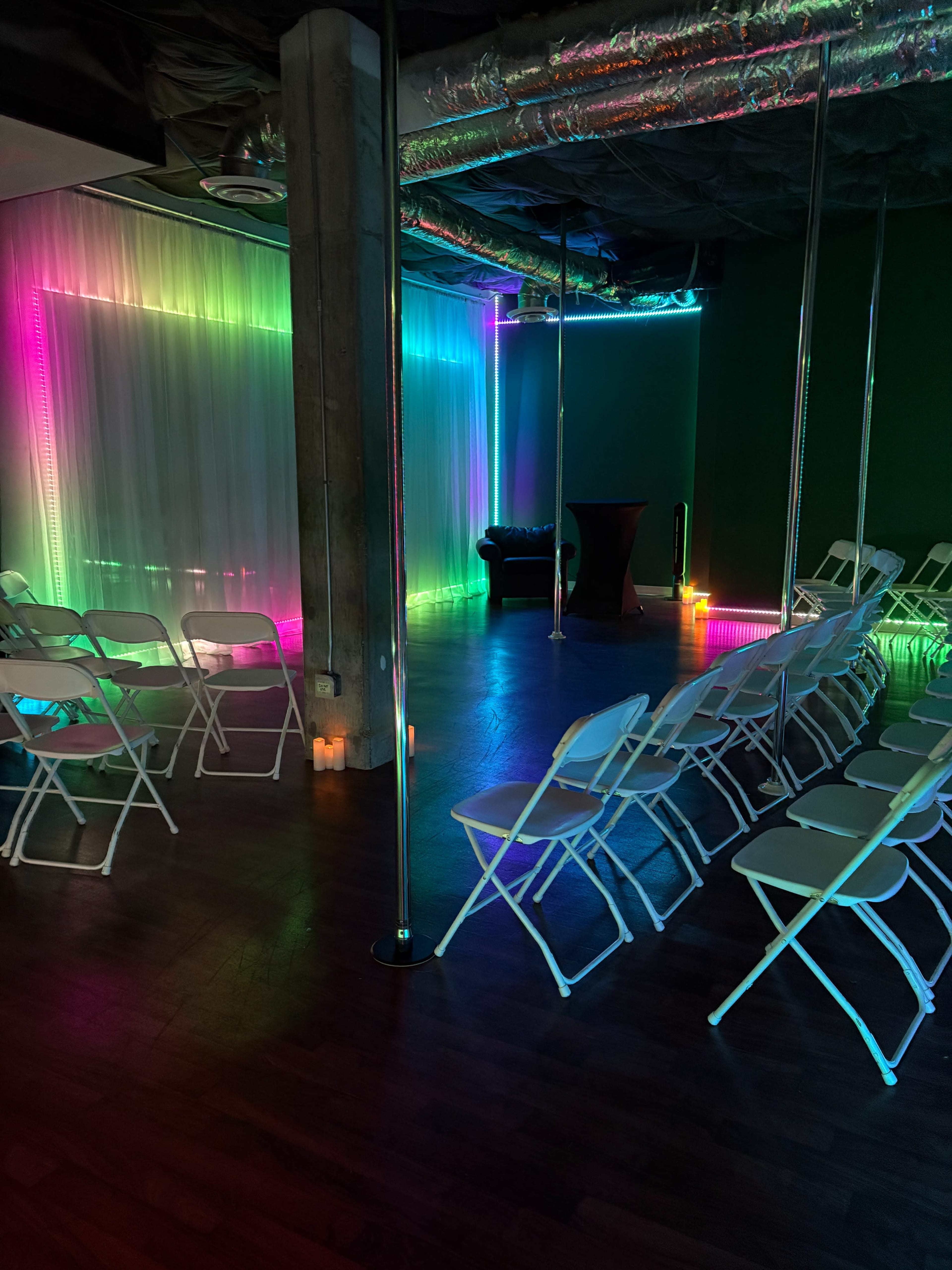The room features folding chairs arranged in rows, illuminated by colorful lights, with a pole on one side and a lounge area in the corner.