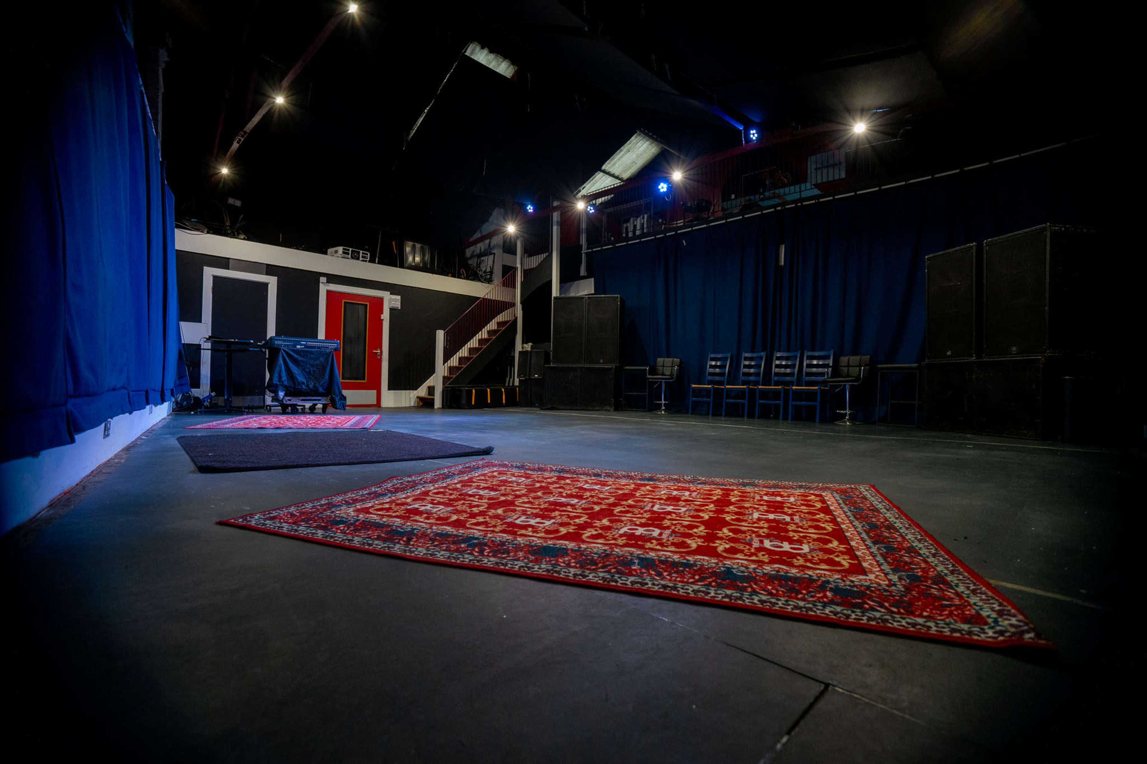 The image shows a performance space with a red carpet on the floor, surrounded by black curtains and chairs, and features a staircase in the background.