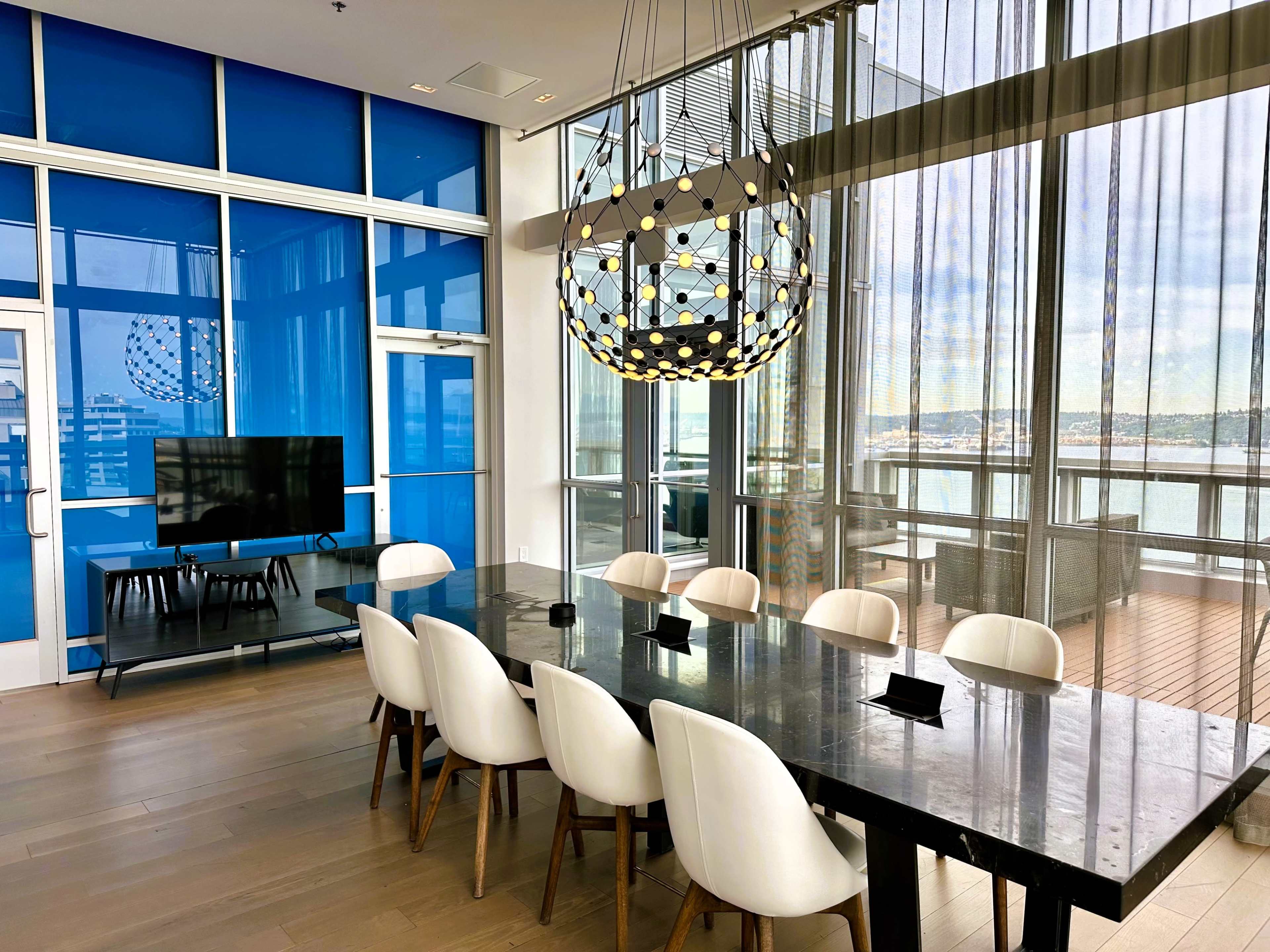 A modern conference room features a long dark table surrounded by white chairs, with large windows and a decorative light fixture overhead.
