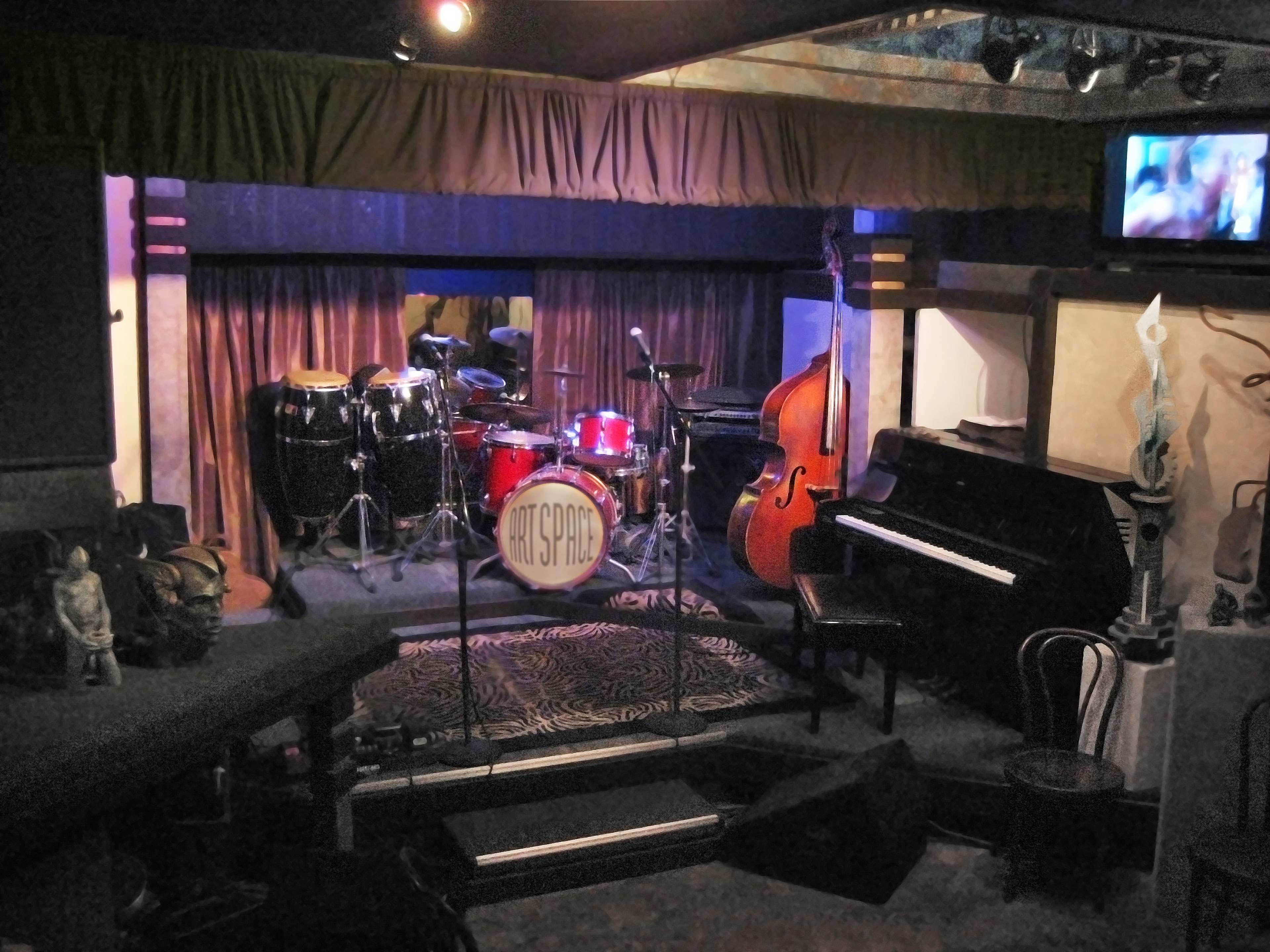 A stage set up for a performance, featuring a drum kit, a double bass, and a piano, with decorative elements in a dimly lit venue.