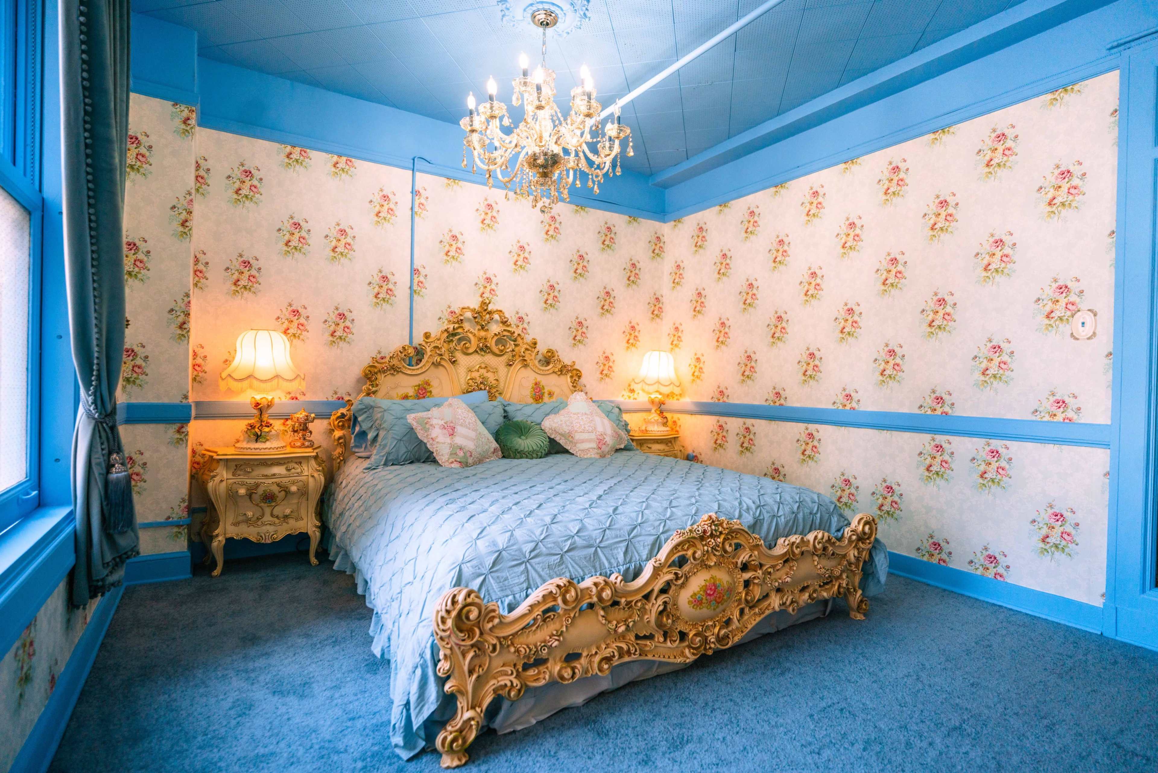 A bedroom with a large, ornate bed, floral wallpaper, a chandelier, and two bedside lamps.