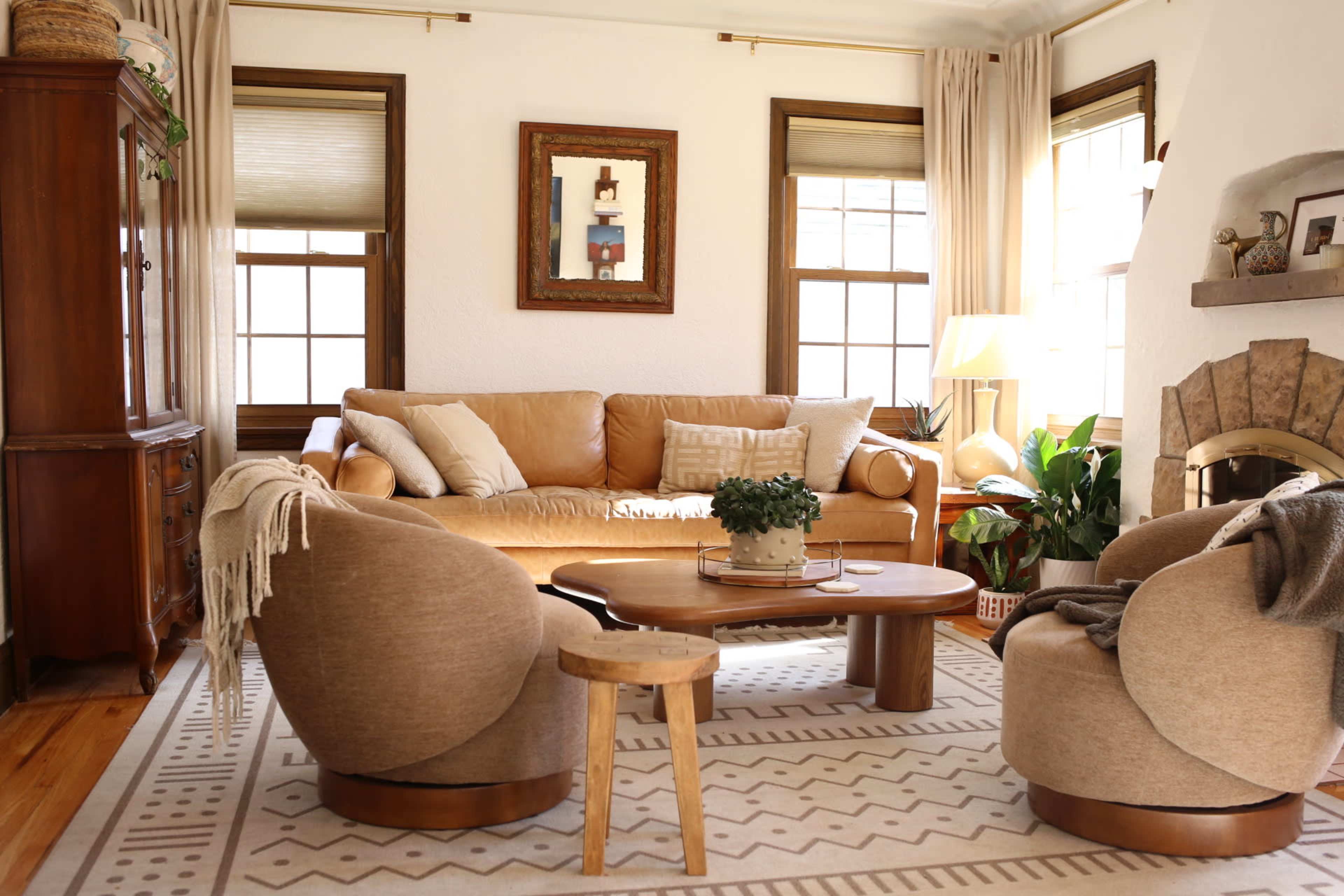 The image shows a cozy living room with a leather sofa, two circular chairs, a wooden coffee table, and decorative plants near large windows.