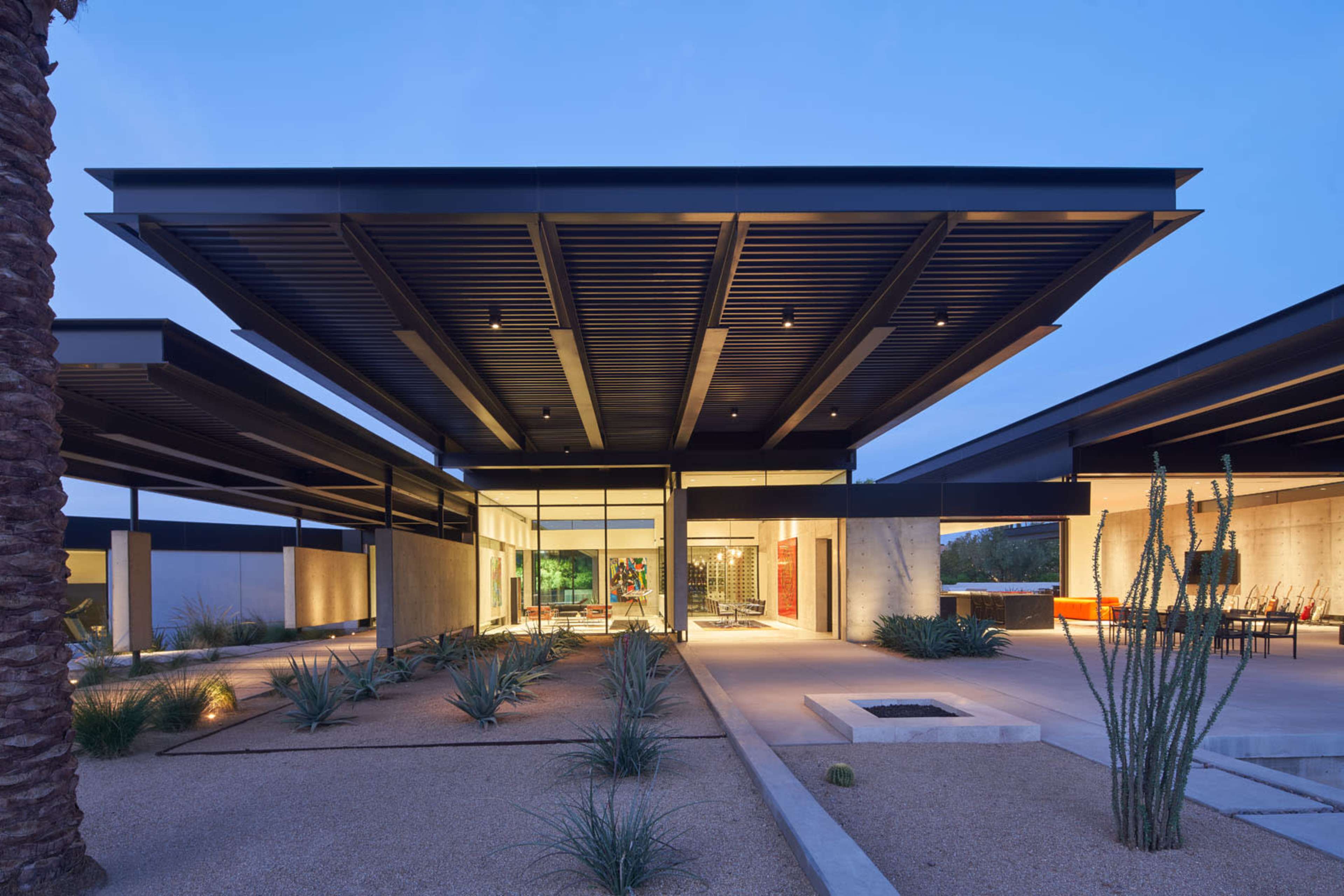 A modern architectural structure with flat roofs, large glass windows, and surrounding desert landscaping, illuminated under twilight.