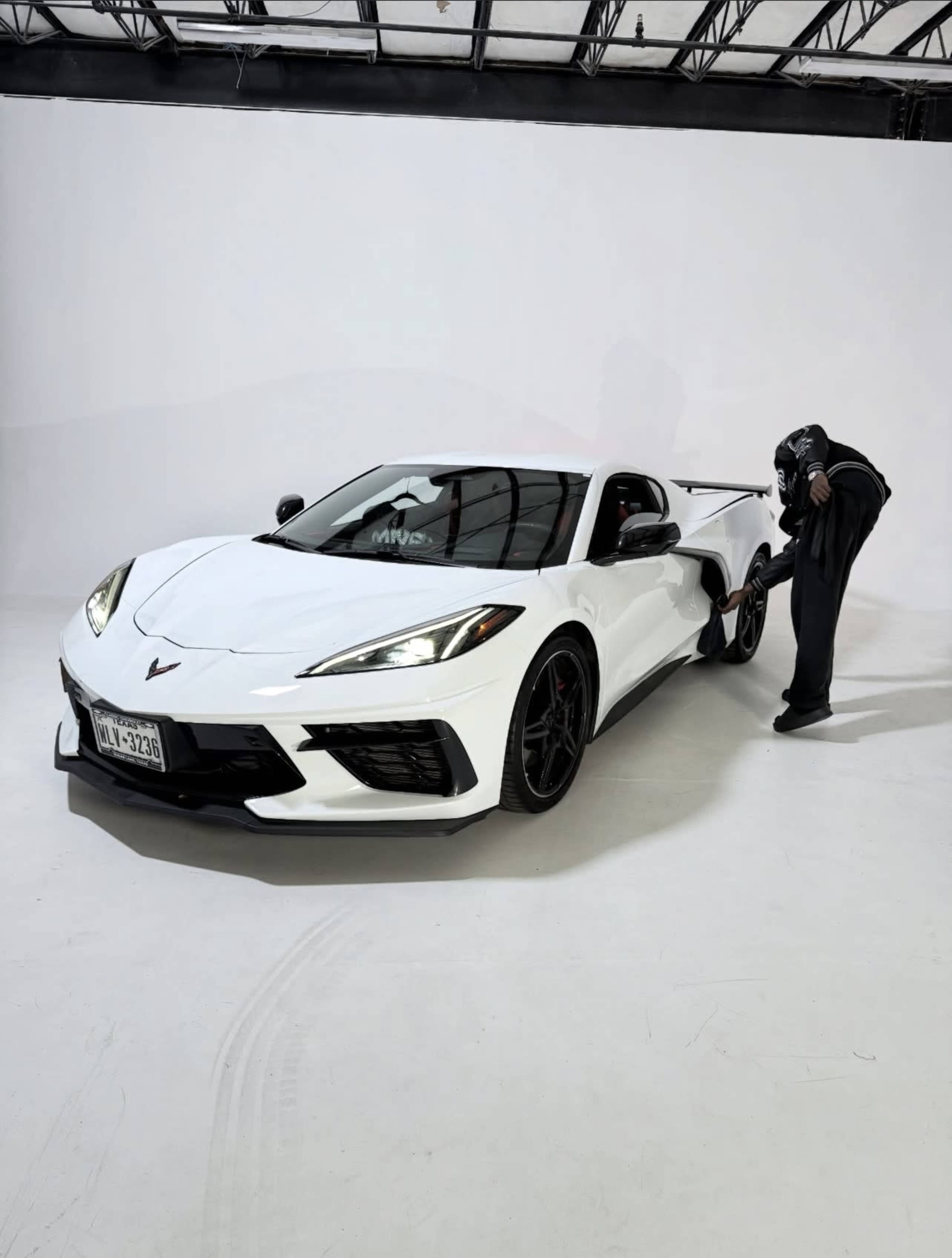 A person in dark clothing stands next to a white Chevrolet Corvette C8 in a studio with a plain background.