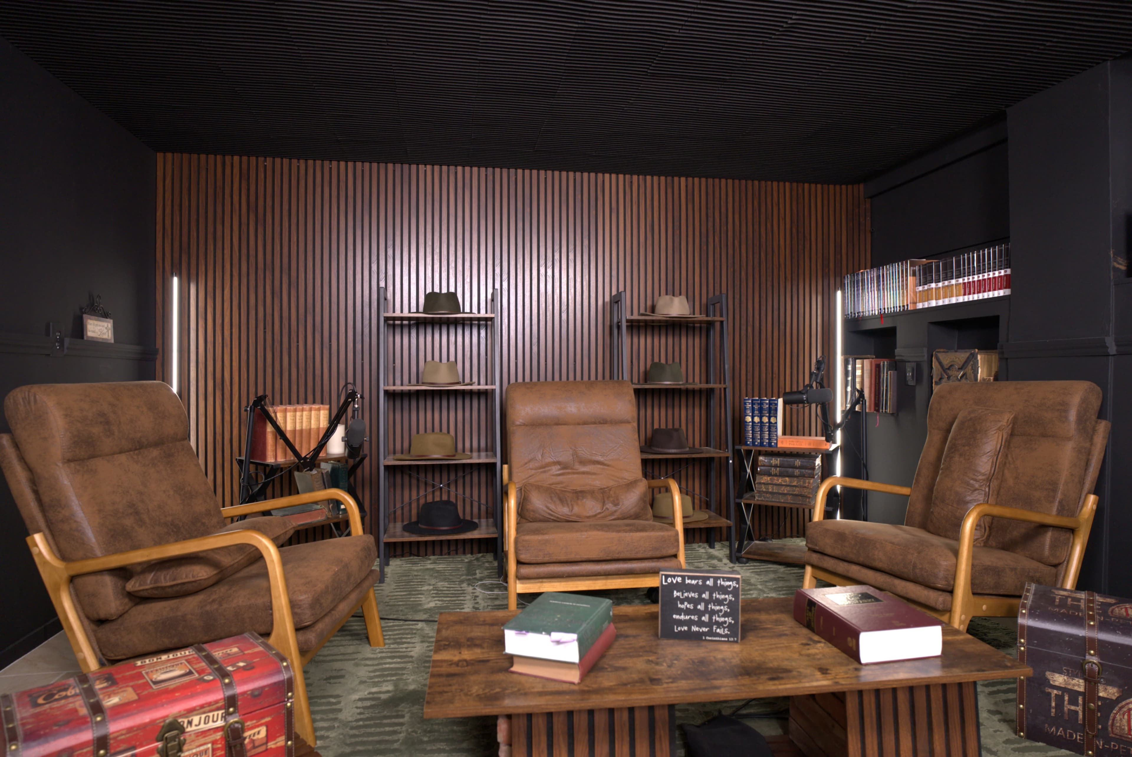 The image shows a cozy interior space with three brown leather armchairs, a wooden coffee table, and shelves lined with hats and books against a wooden-paneled wall.