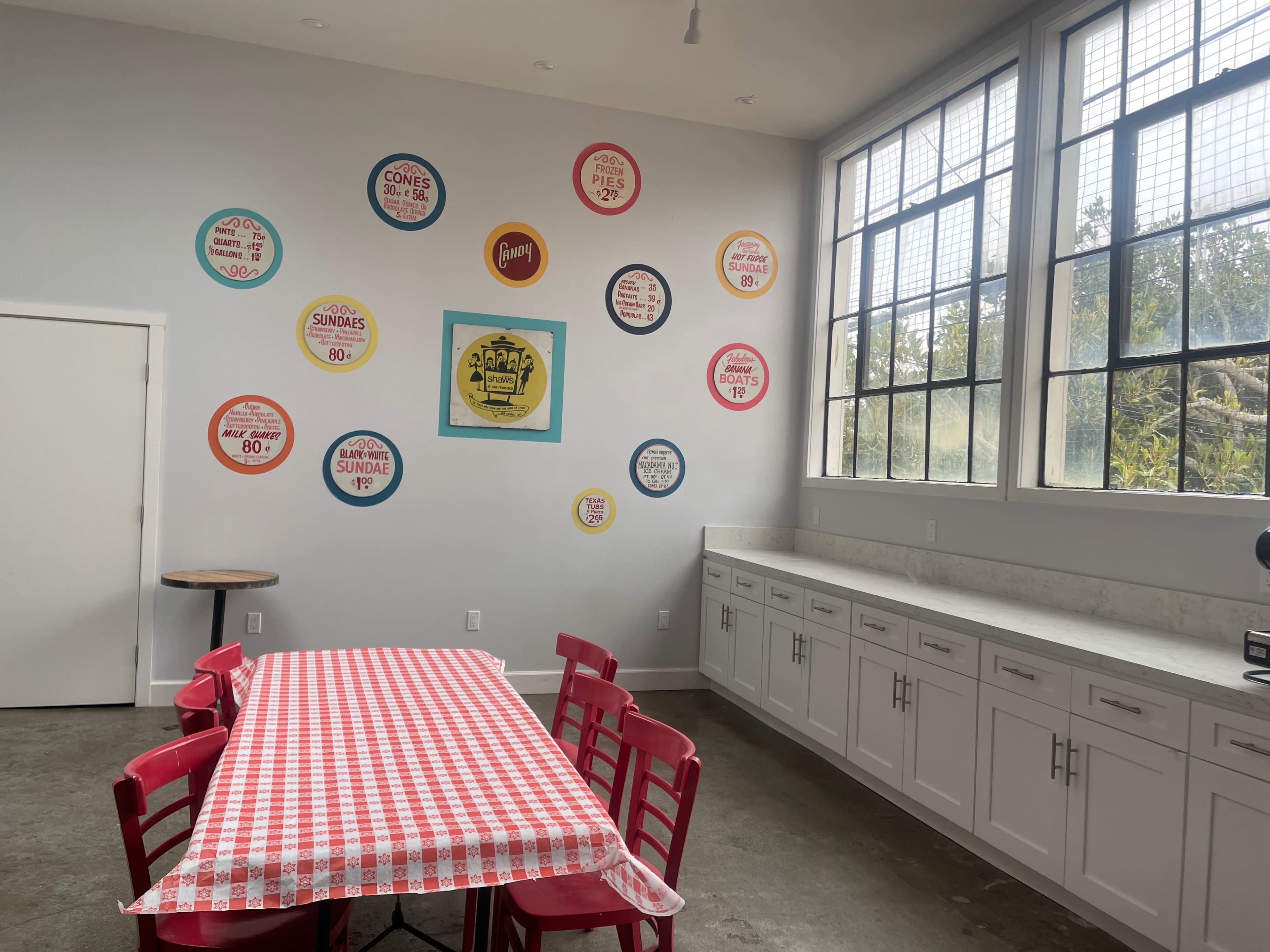 A bright room features a red-checkered table surrounded by red chairs, with vintage-style signs displayed on the wall.