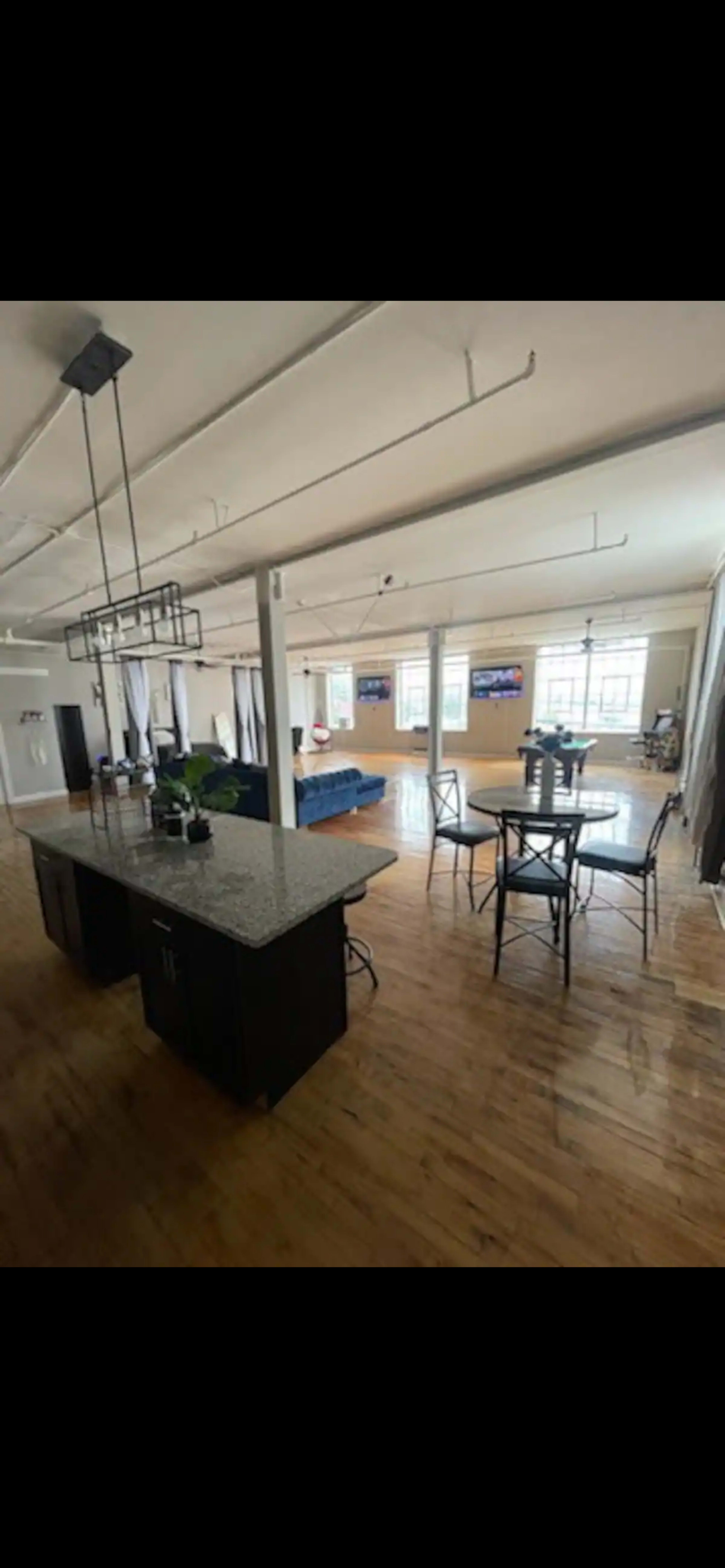 The image depicts a spacious, modern interior with a kitchen island, dining area, and multiple seating spaces, illuminated by natural light from large windows.