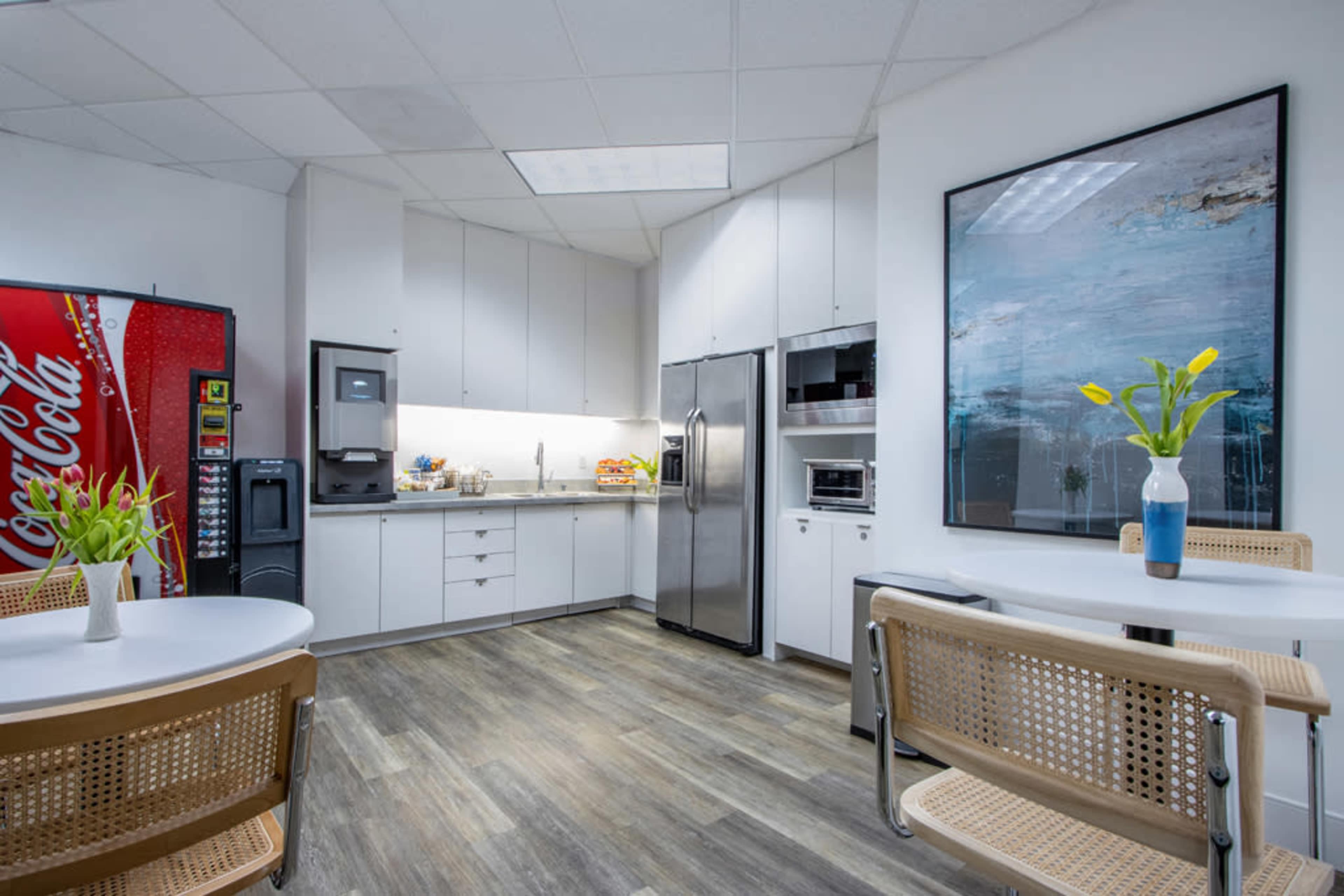 The image shows a modern kitchen area with stainless steel appliances, including a refrigerator and microwave, surrounded by a table and chairs, along with a vending machine and water dispenser.