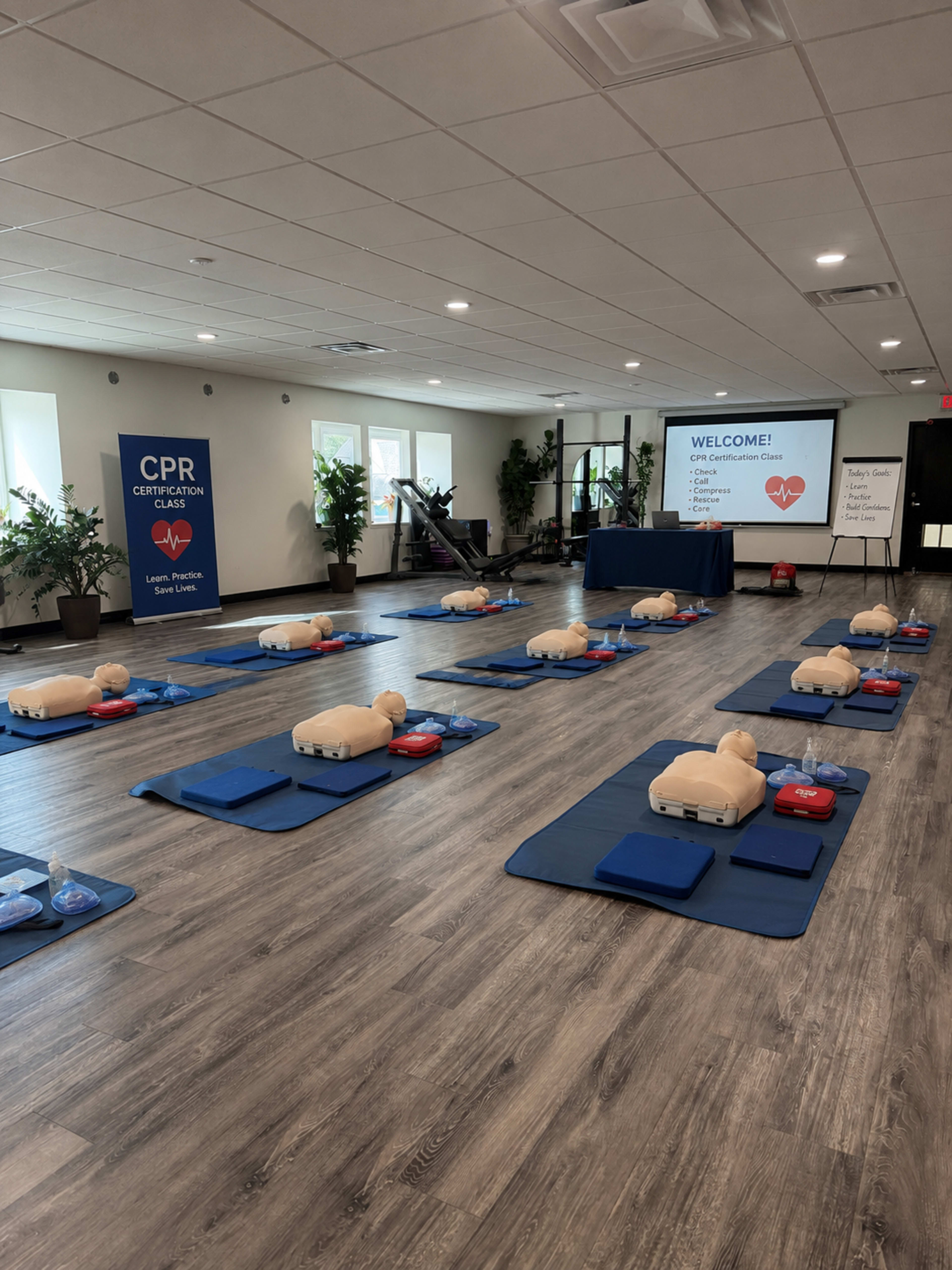 The image shows a CPR certification class setup with multiple training mannequins arranged on blue mats, and a large screen displaying welcome messages in a well-lit room.