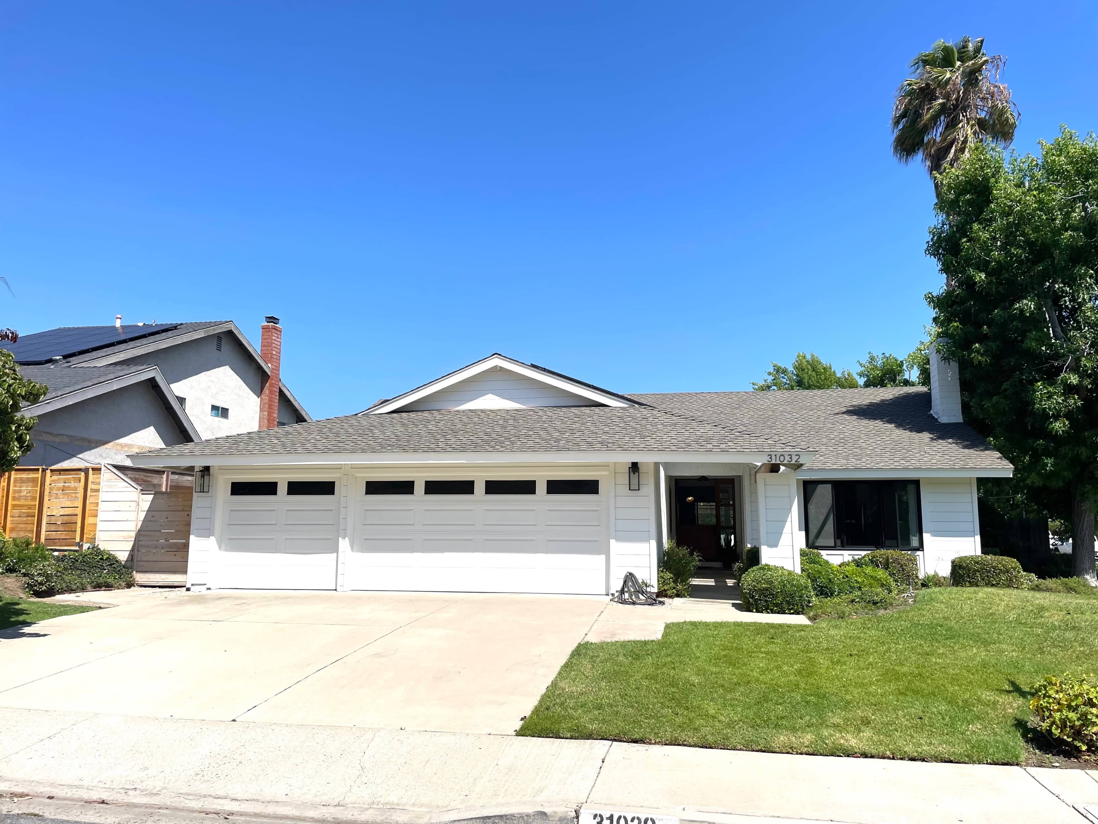 A single-story house with a gray roof and three garage doors is set on a green lawn under a clear blue sky.