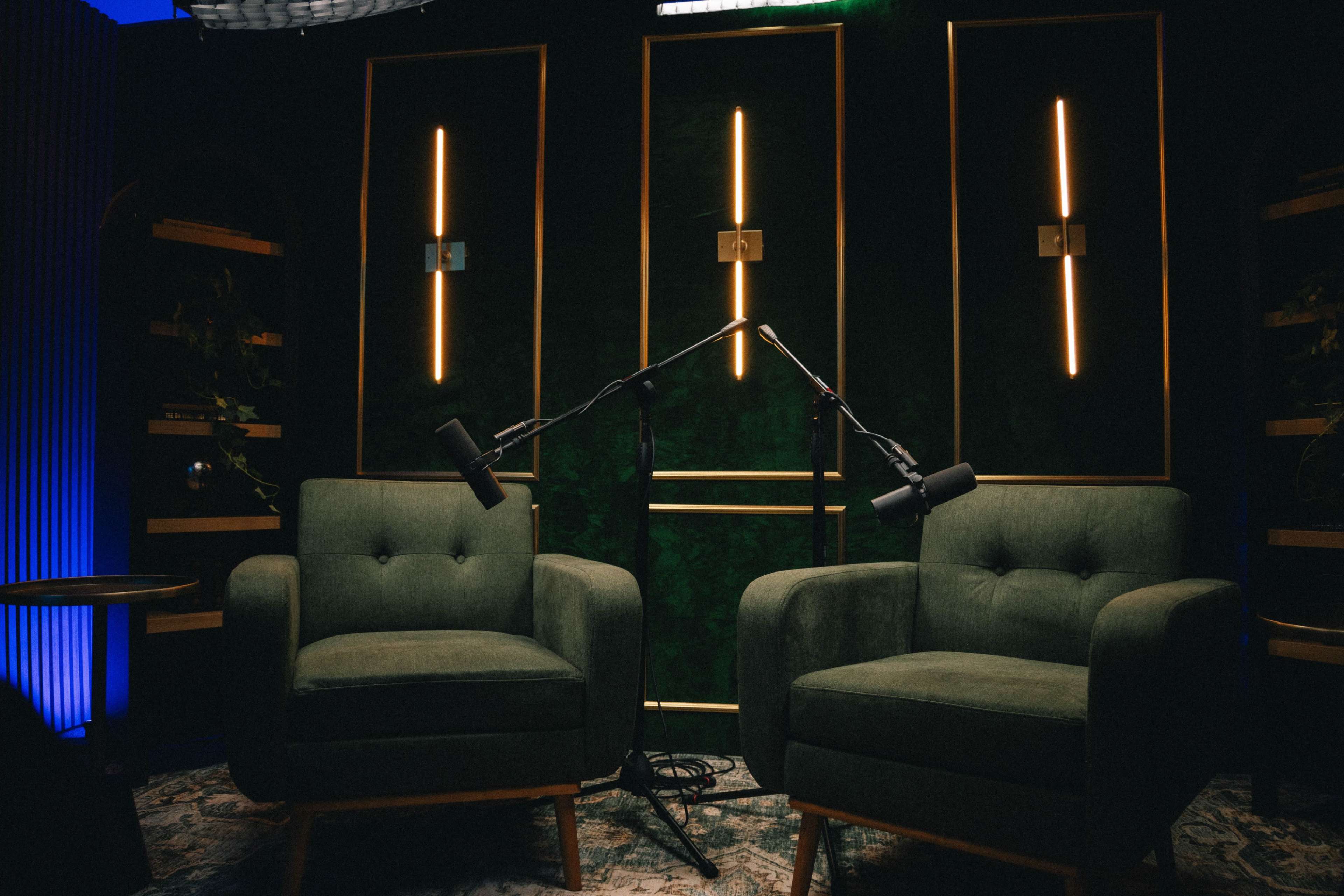 Two green upholstered armchairs are positioned opposite each other in a dimly lit room, with microphones mounted on stands above them and illuminated vertical light fixtures in the background.