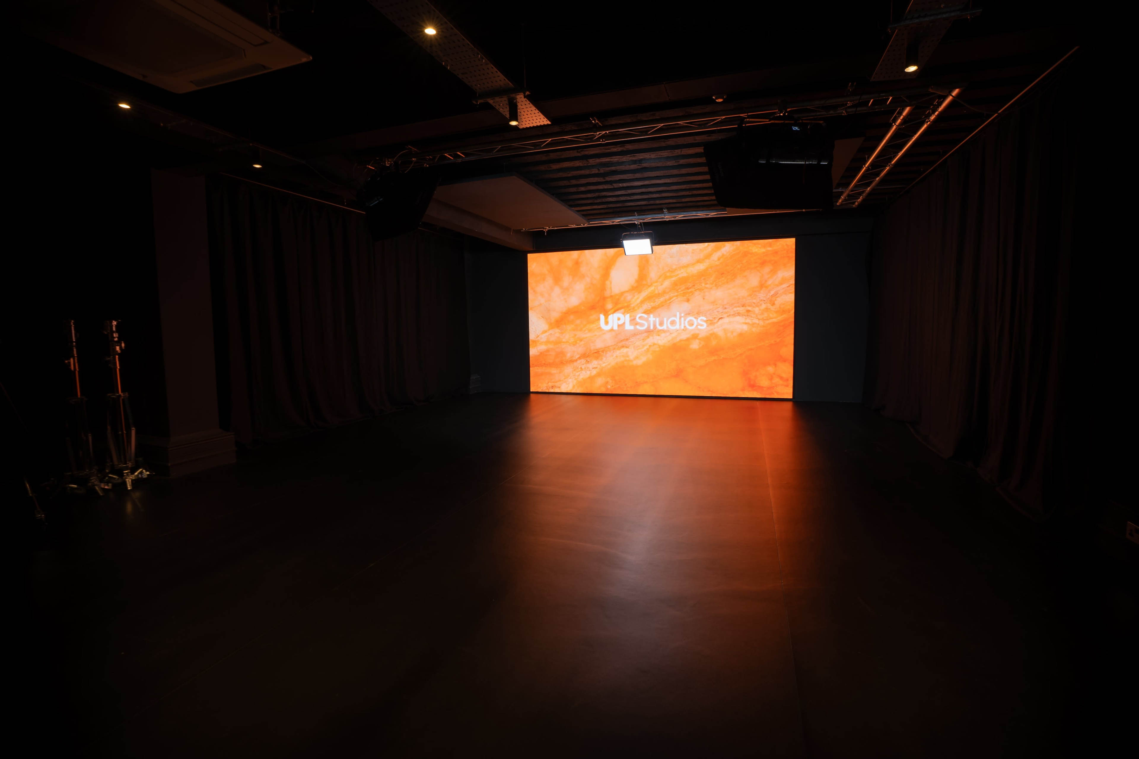 The image shows a dimly lit studio space with a large screen displaying the logo "UPL Studios" against an orange backdrop.
