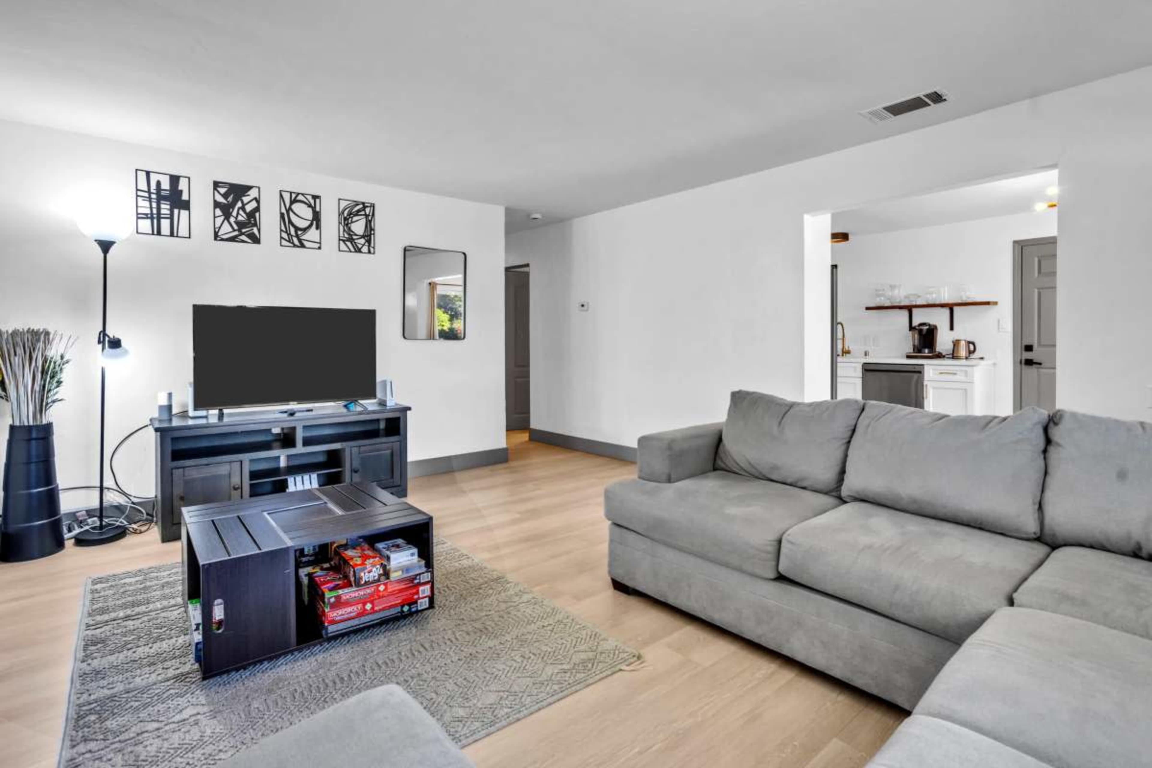 The image shows a modern living room with a gray sectional sofa, a coffee table displaying games, and a television on a black media console, alongside a lamp and a doorway leading to a kitchen area.