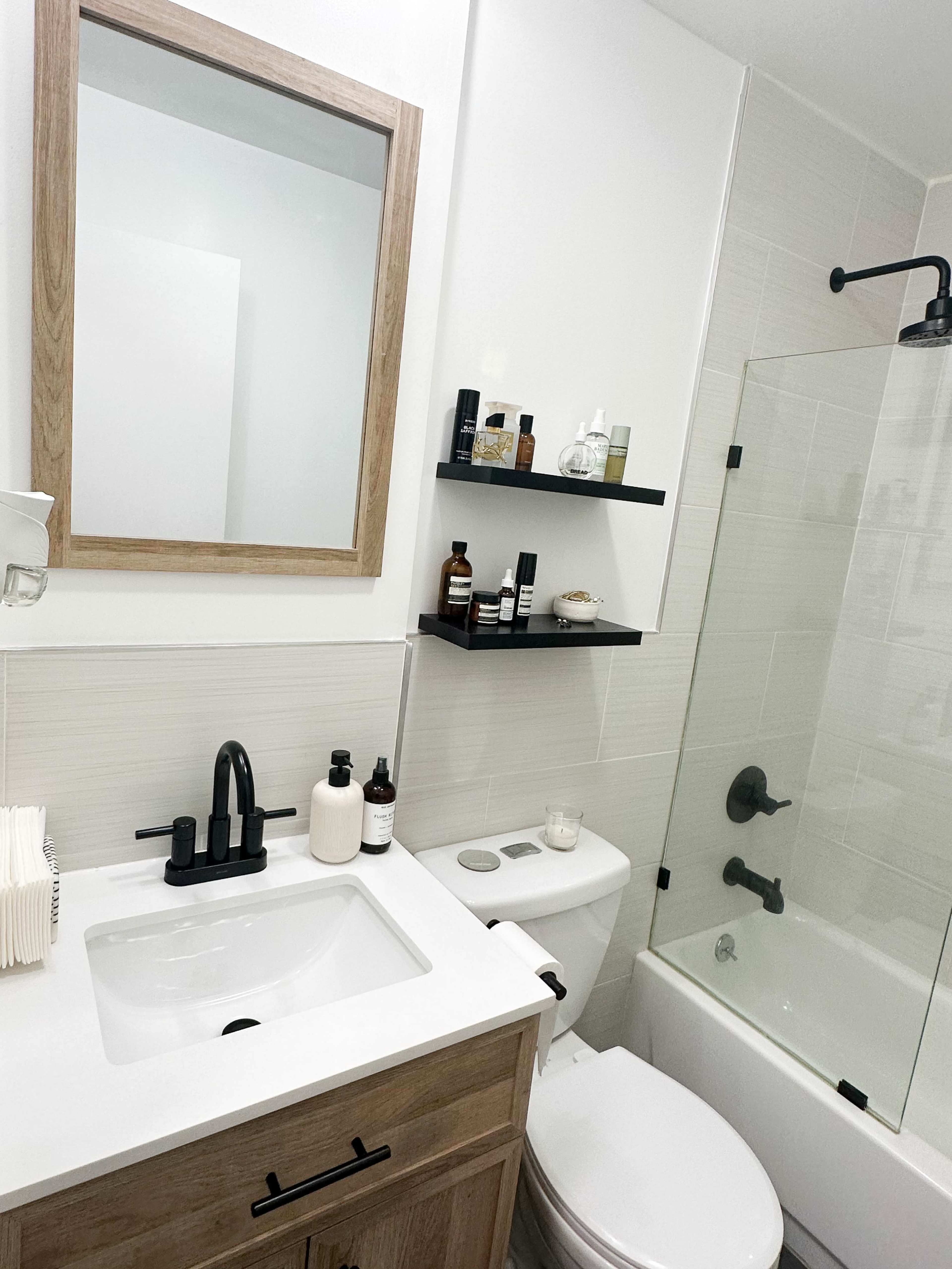 A modern bathroom featuring a white sink with a wooden cabinet, a mirror, a toilet, a glass shower, and open shelves with various skincare products.