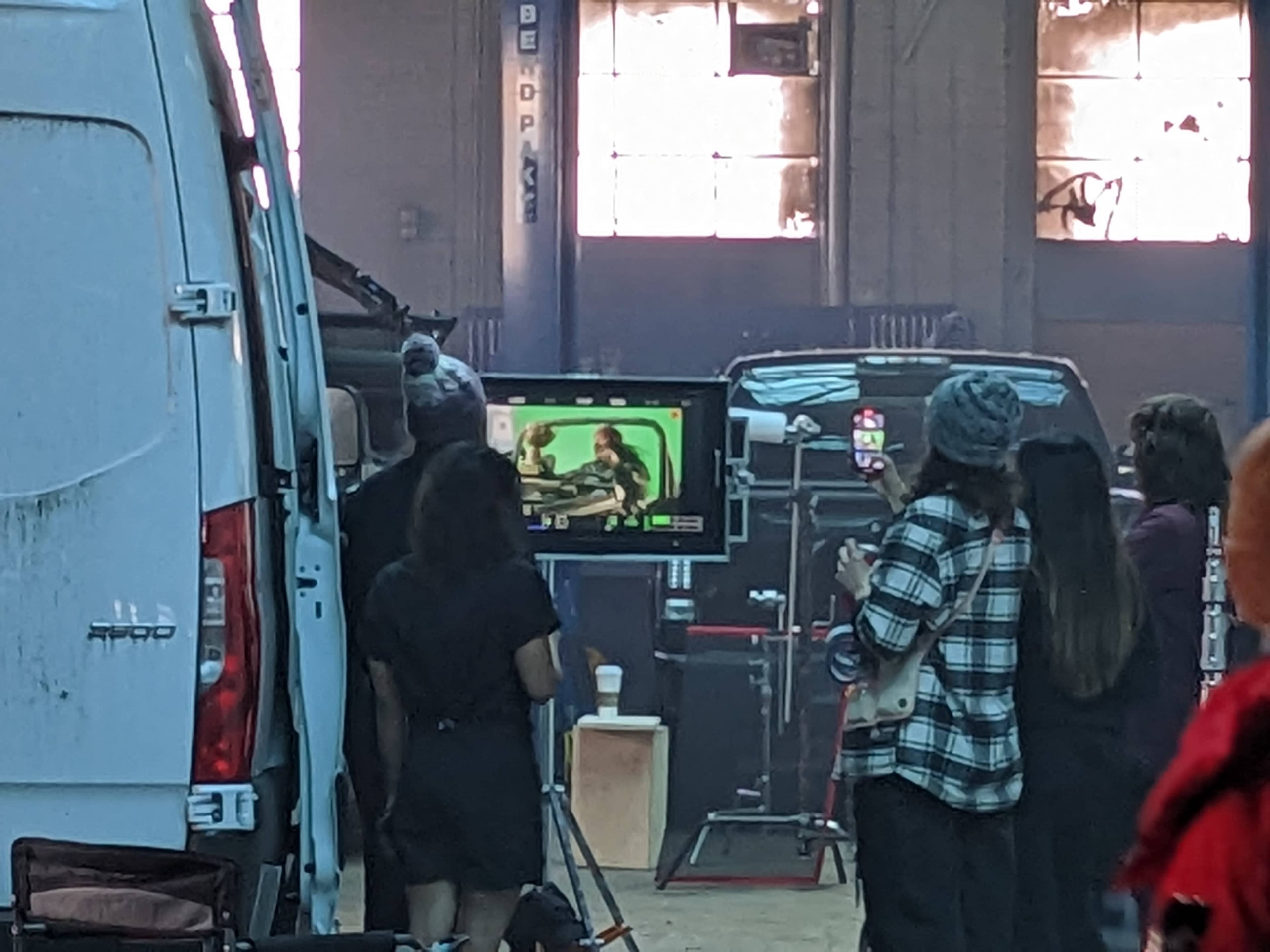 A group of people watches a scene being filmed on a monitor inside a warehouse.