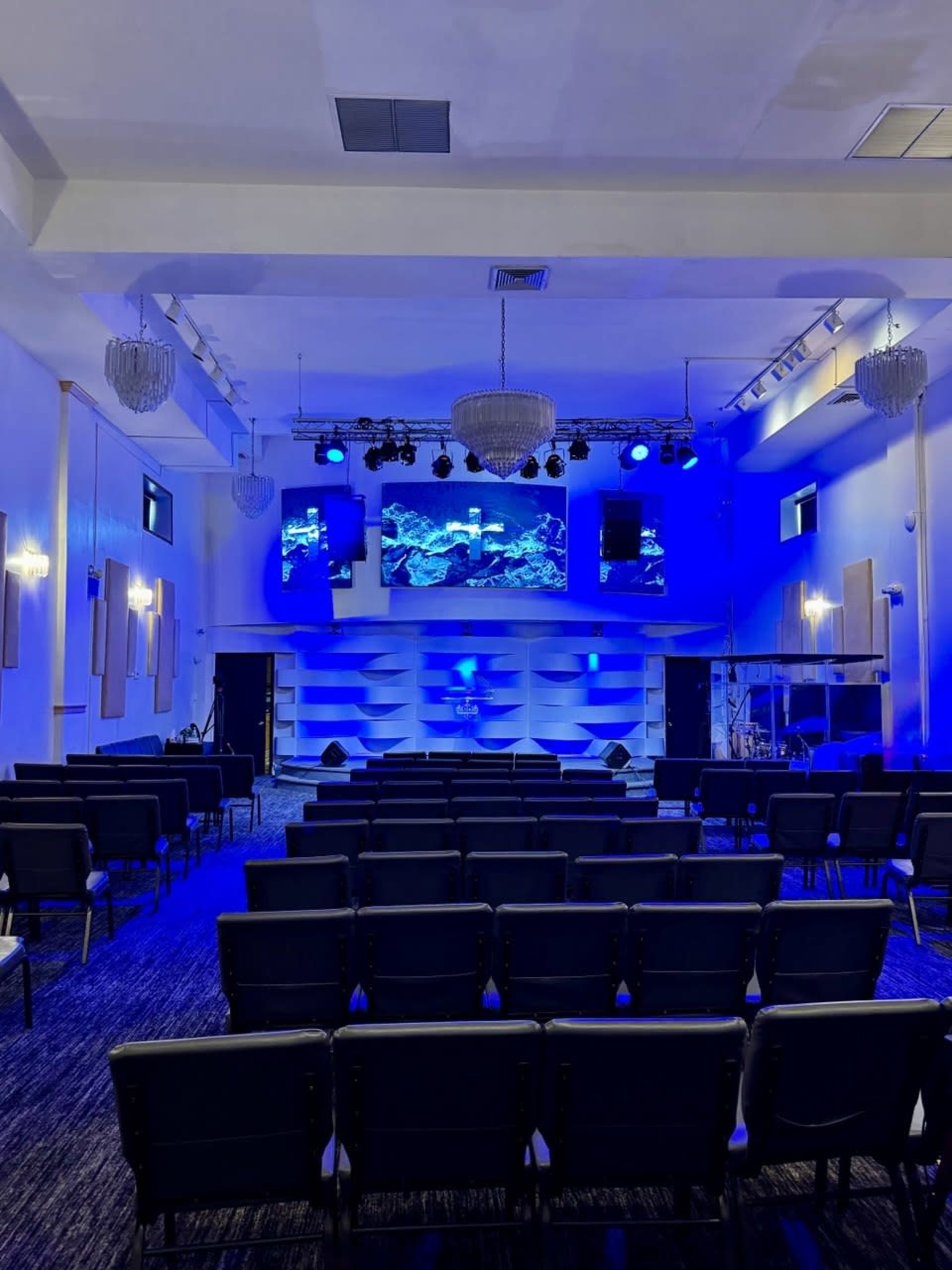 The image shows a modern church interior with rows of black chairs facing a stage illuminated by blue lighting and featuring screens displaying aquatic imagery.