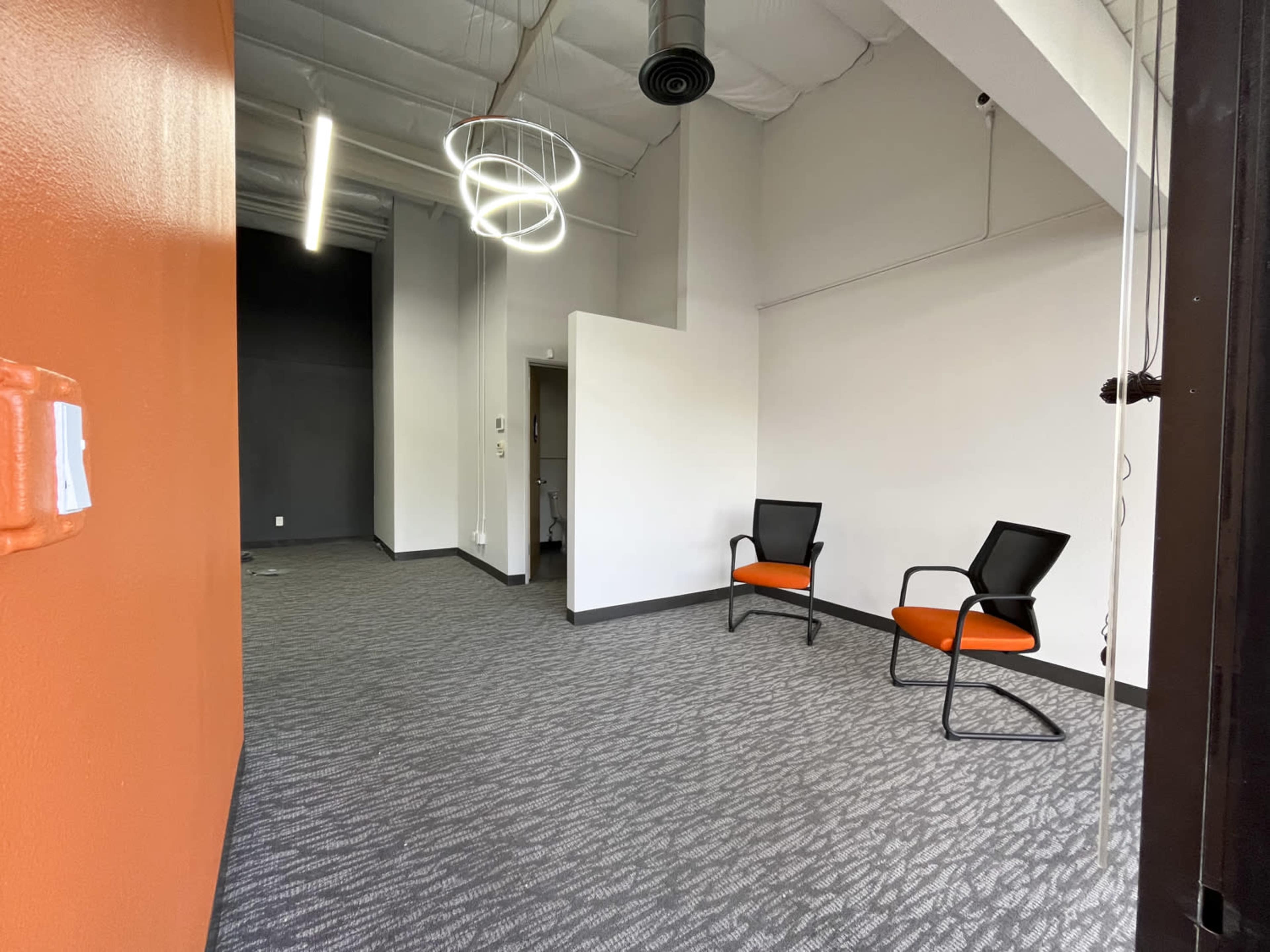 A vacant office space with orange and gray walls, featuring two black chairs on a patterned carpet.