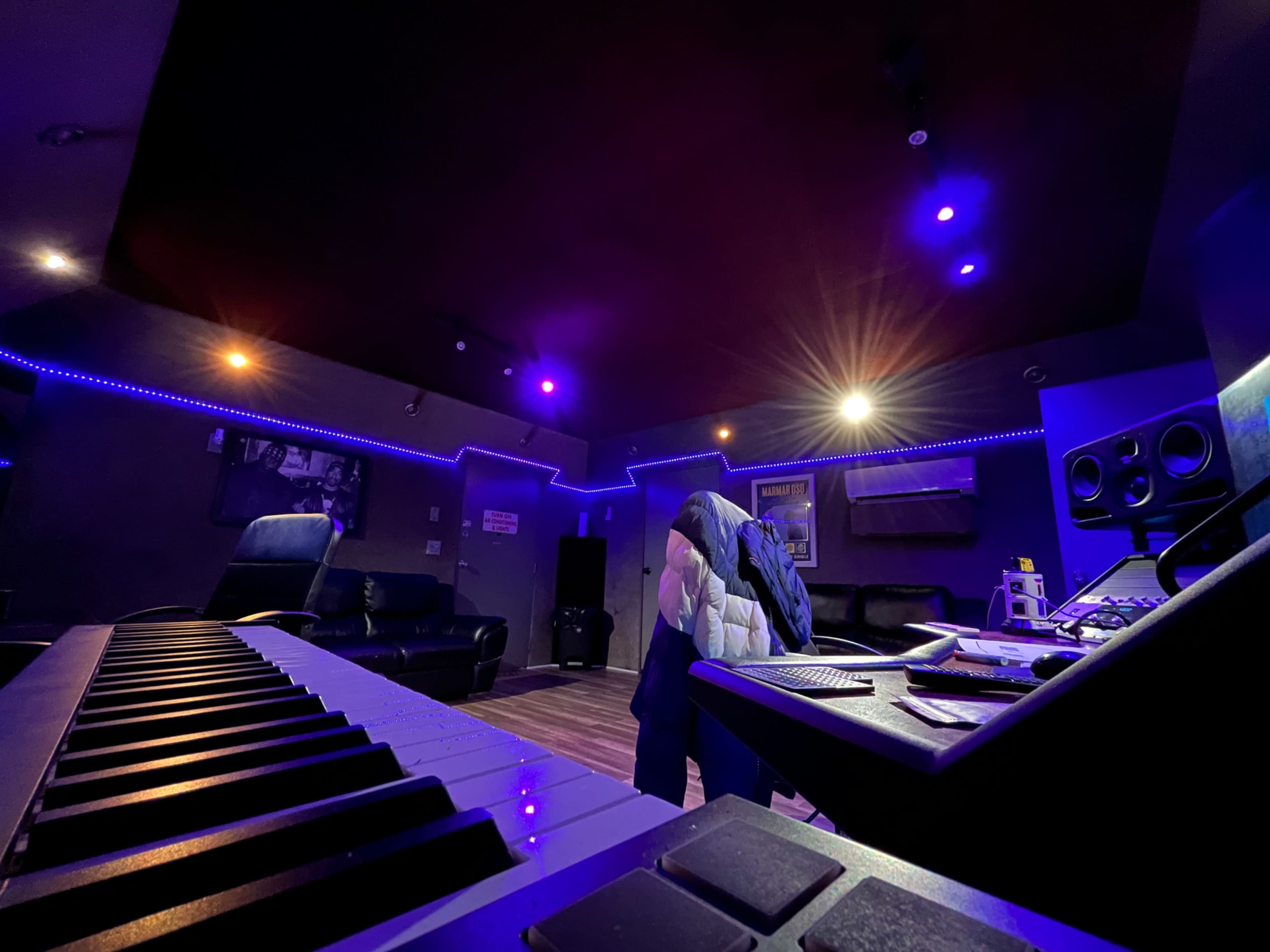 A dimly lit recording studio features a keyboard in the foreground, a chair draped with a jacket, and ambient lighting along the walls.