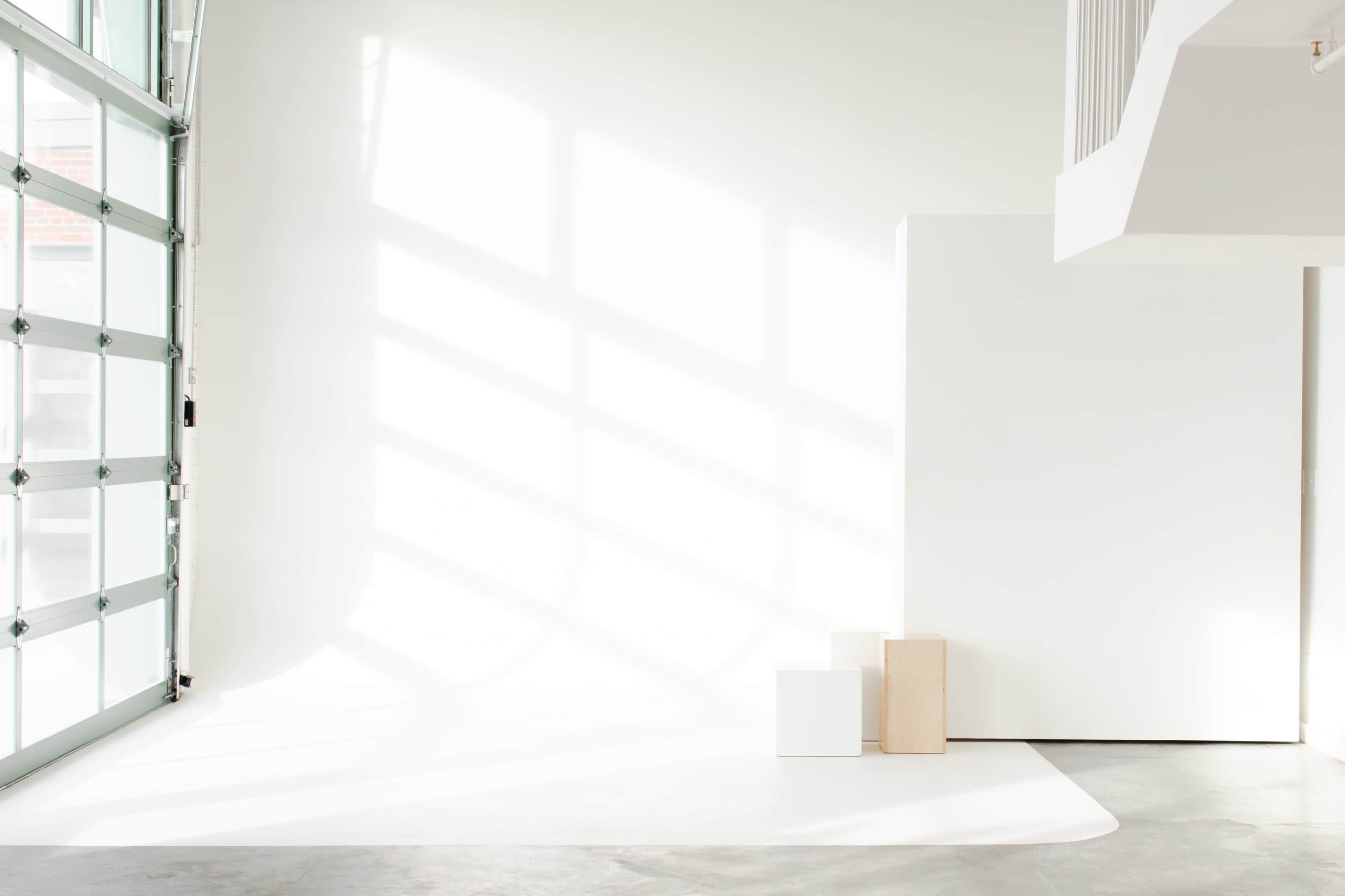 A minimalist interior space with a white backdrop, two wooden blocks, and large windows casting geometric shadows on the floor.