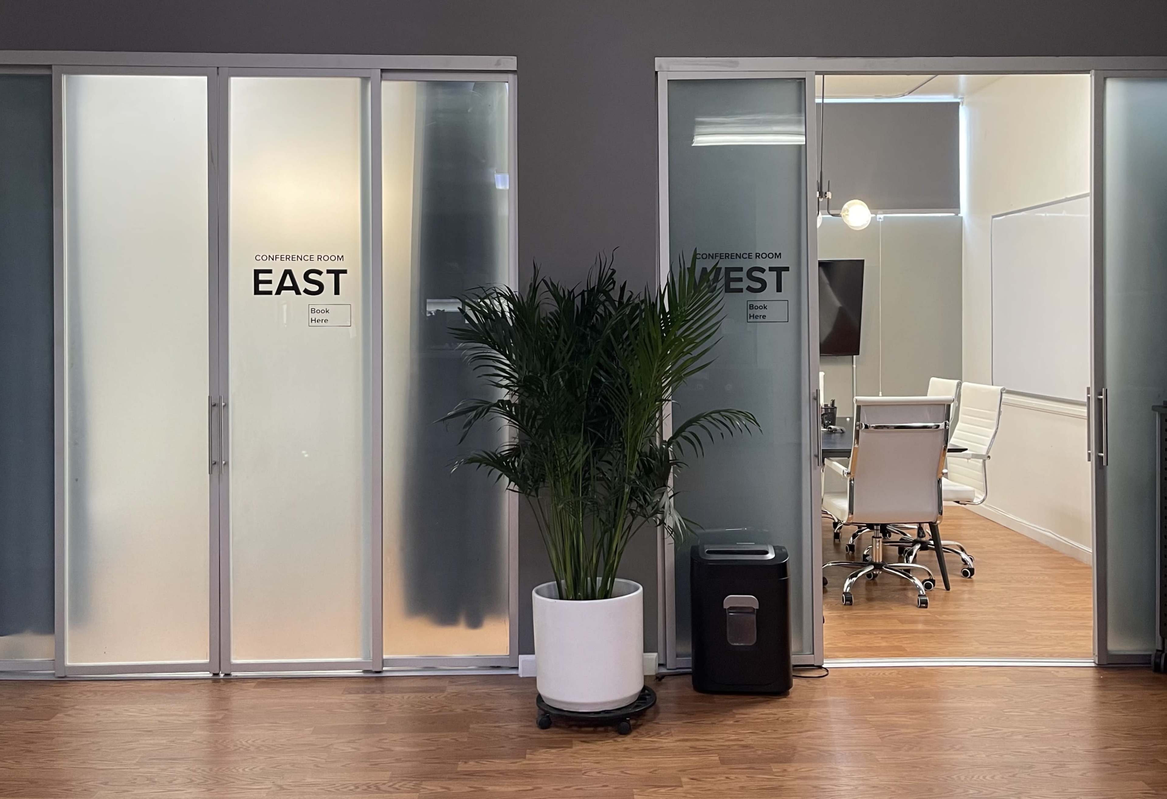 Two glass doors labeled "EAST" and "WEST" leading to conference rooms, with a potted plant and a trash can in the foreground.