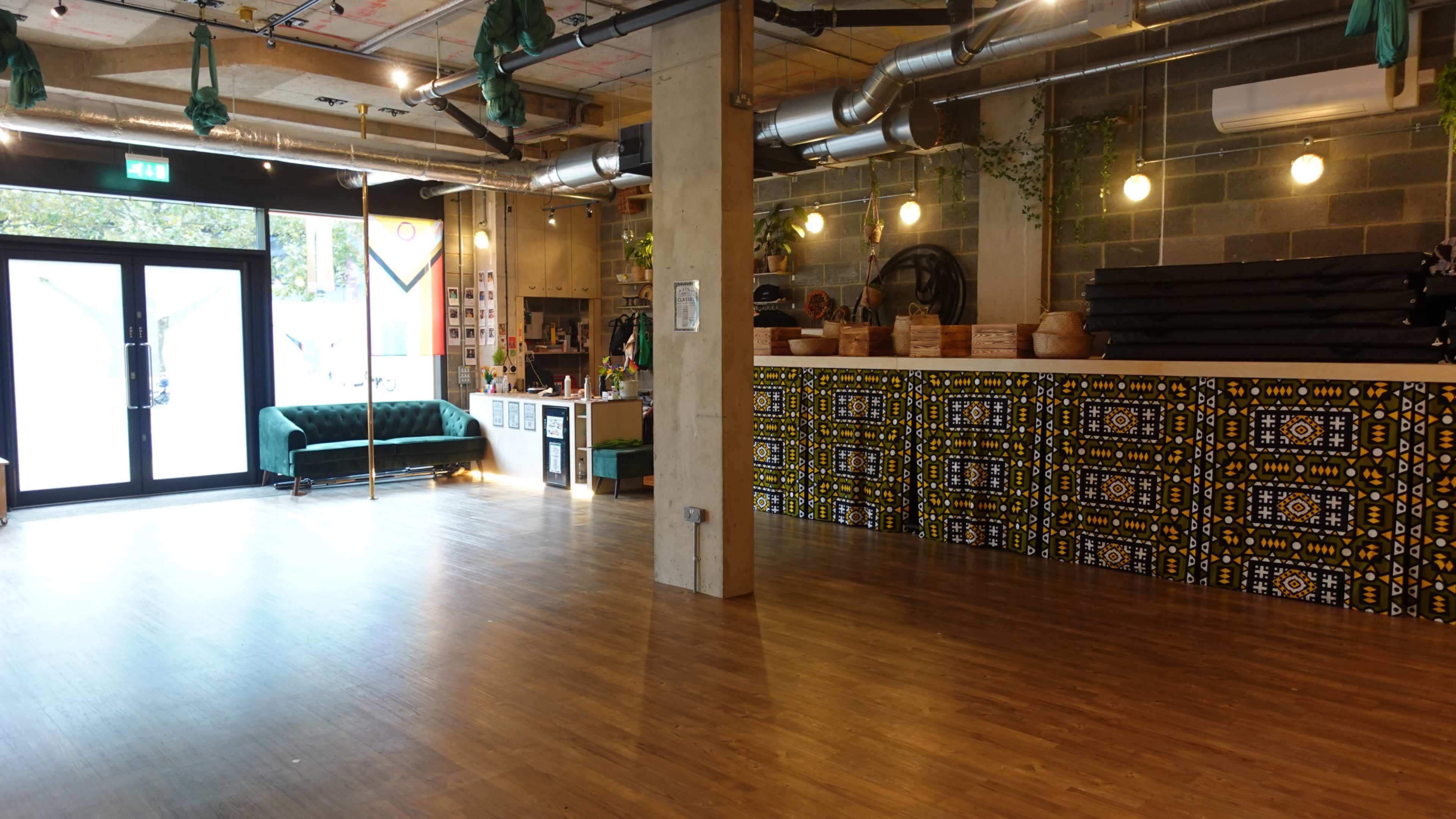 The image shows a modern indoor space with wooden flooring, featuring a green upholstered sofa, a reception area with colorful patterned tiles, and large windows allowing natural light to enter.