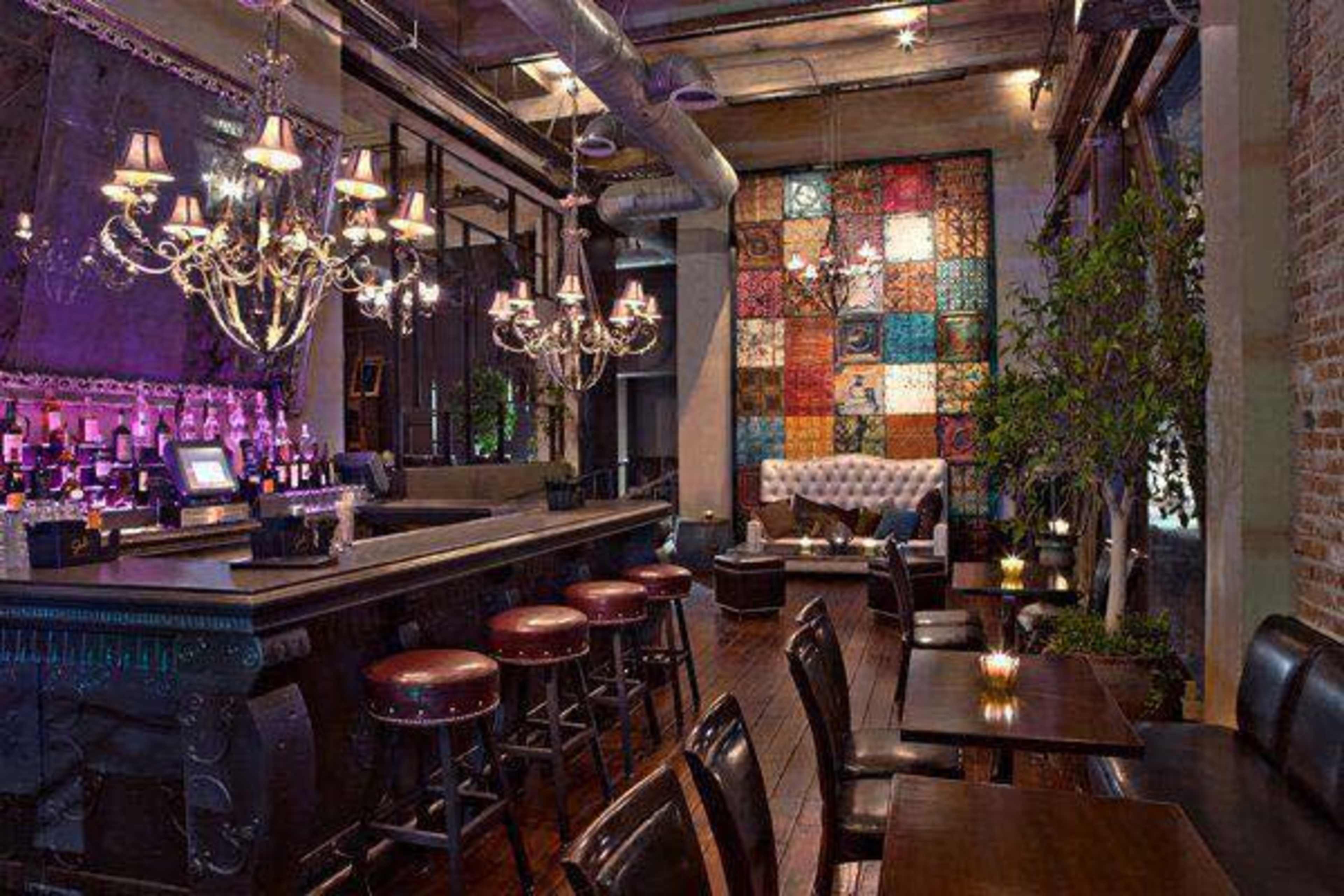 A dimly lit bar with a polished counter, high stools, and decorative chandeliers, complemented by a colorful wall art piece and a lounge area with a tufted sofa.