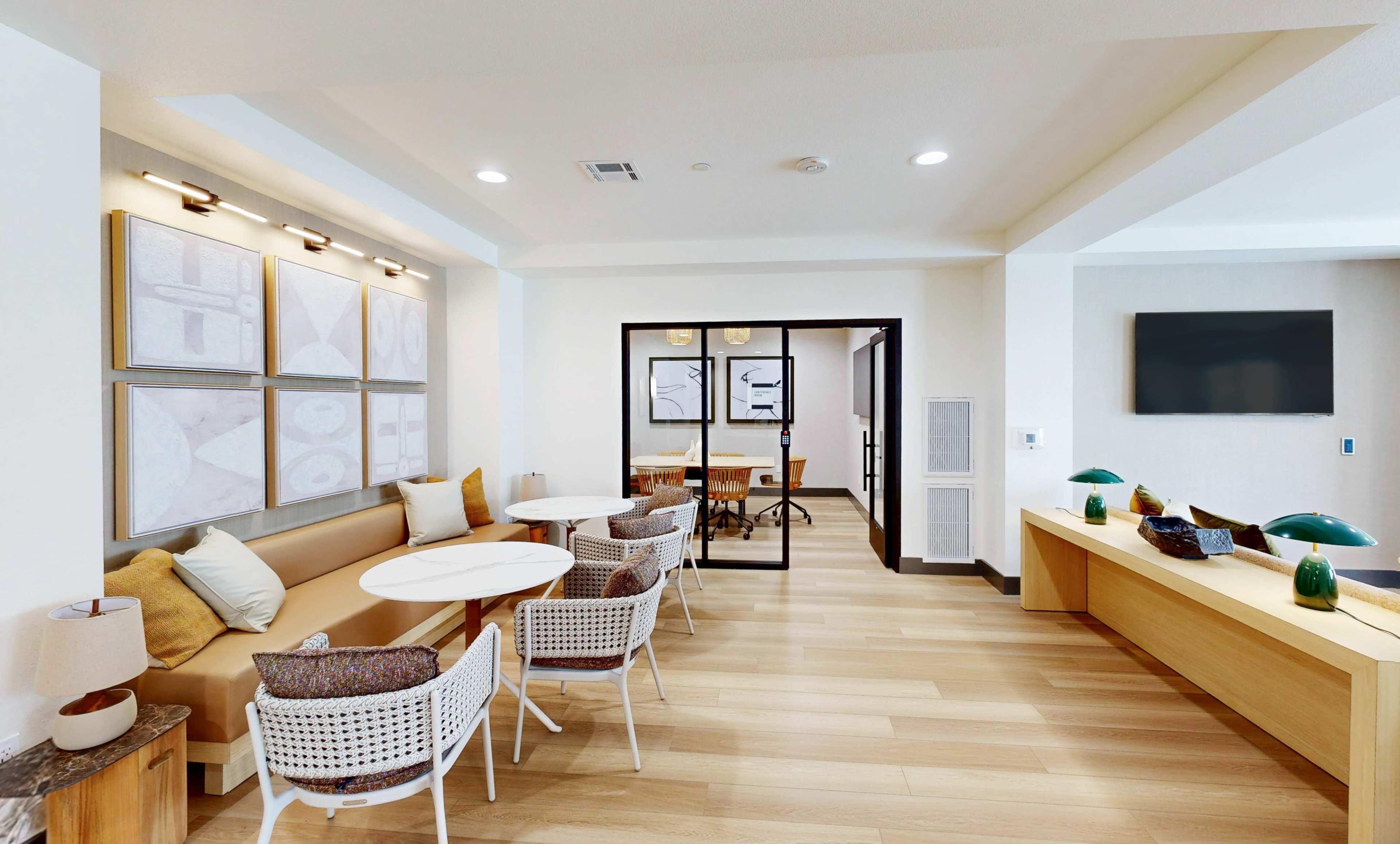 The image shows a modern interior of a room featuring a seating area with several round tables, chairs, and wall art, leading to a glass door entryway into another space.