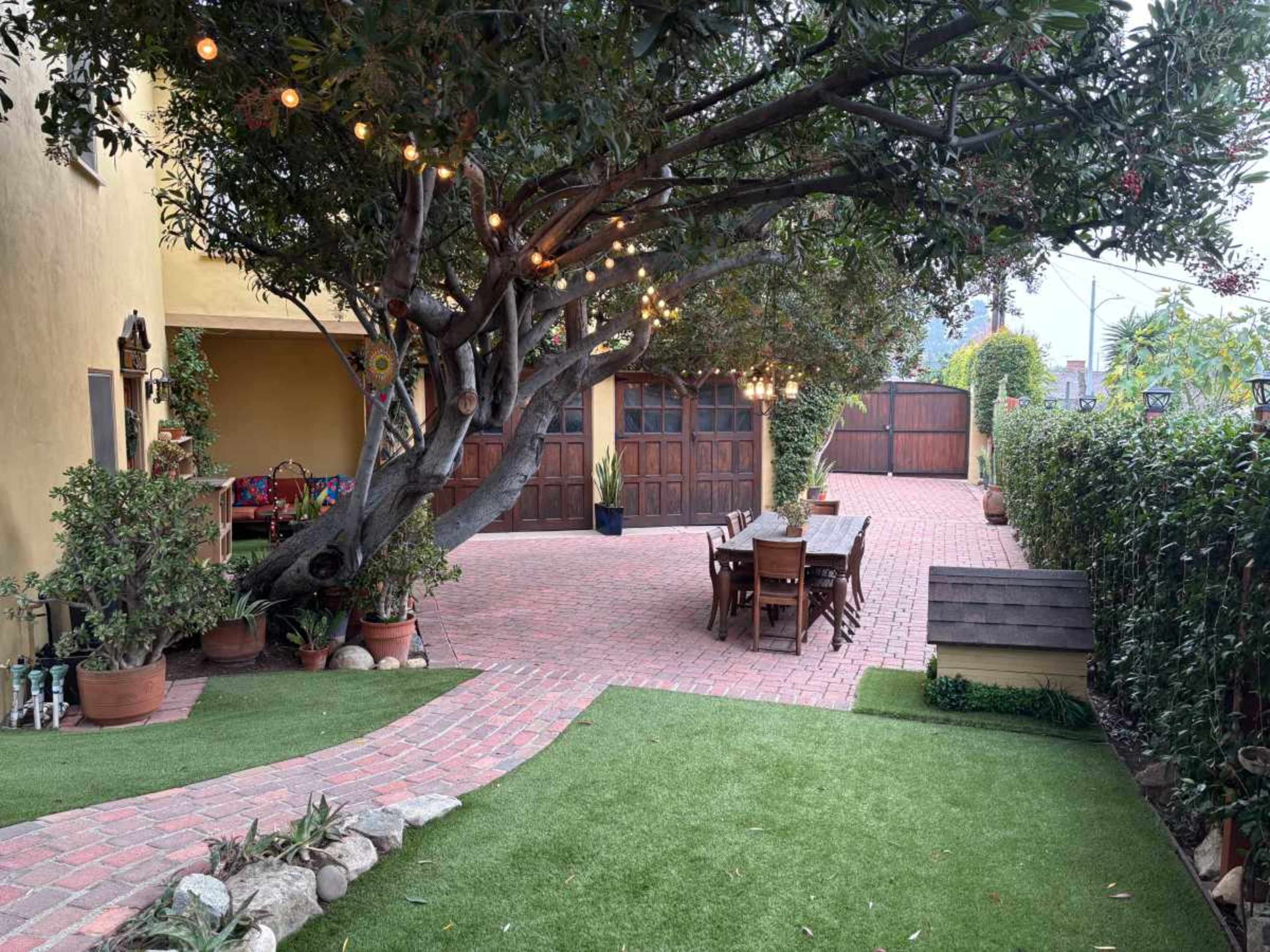 The image shows a spacious outdoor area with a brick patio, wooden furniture under a large tree adorned with lights, and a gated entrance in the background.