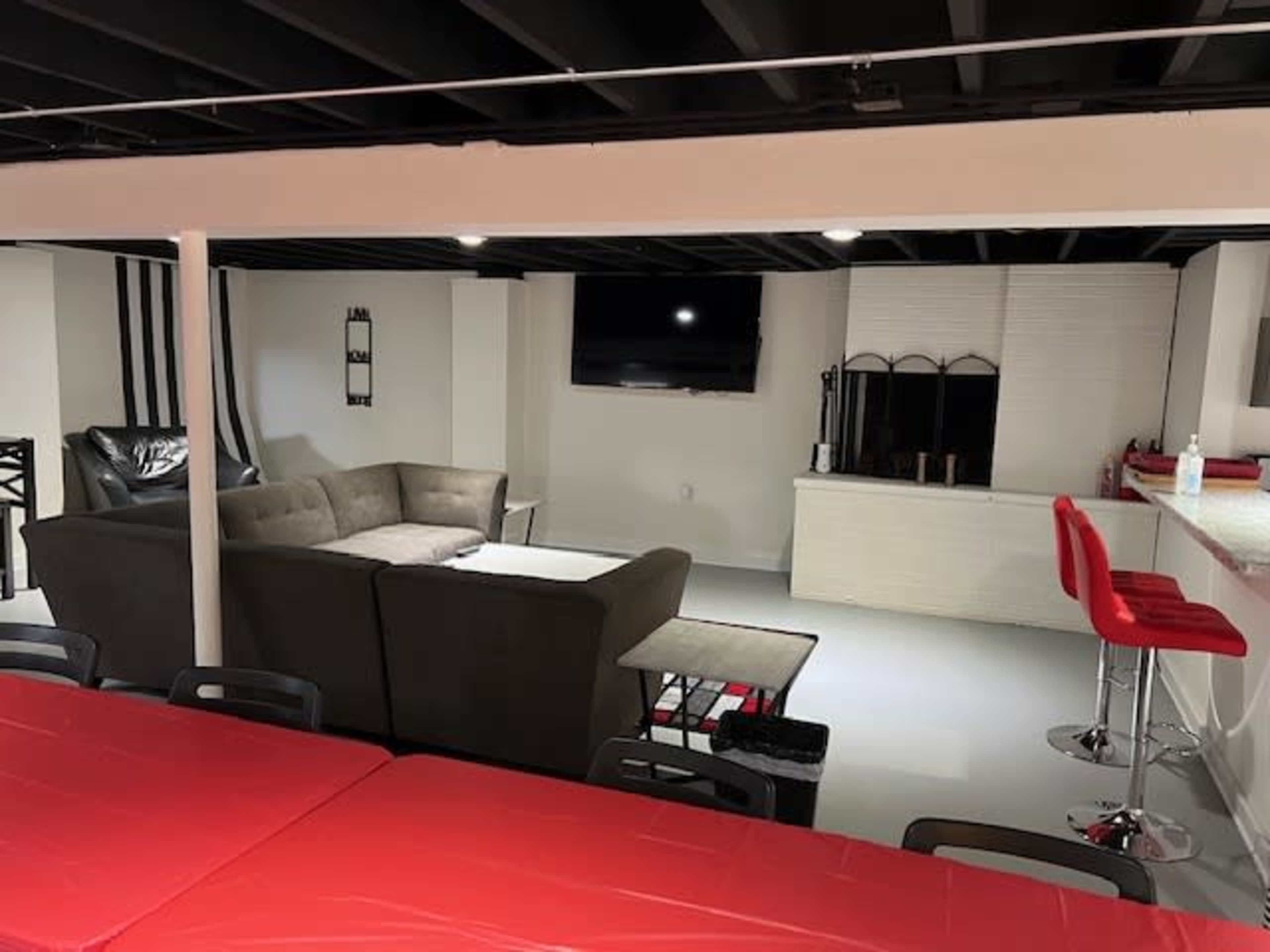 A modern basement living area with a sectional sofa, a television mounted on the wall, and a dining table with red tablecloths, along with barstools at a counter.