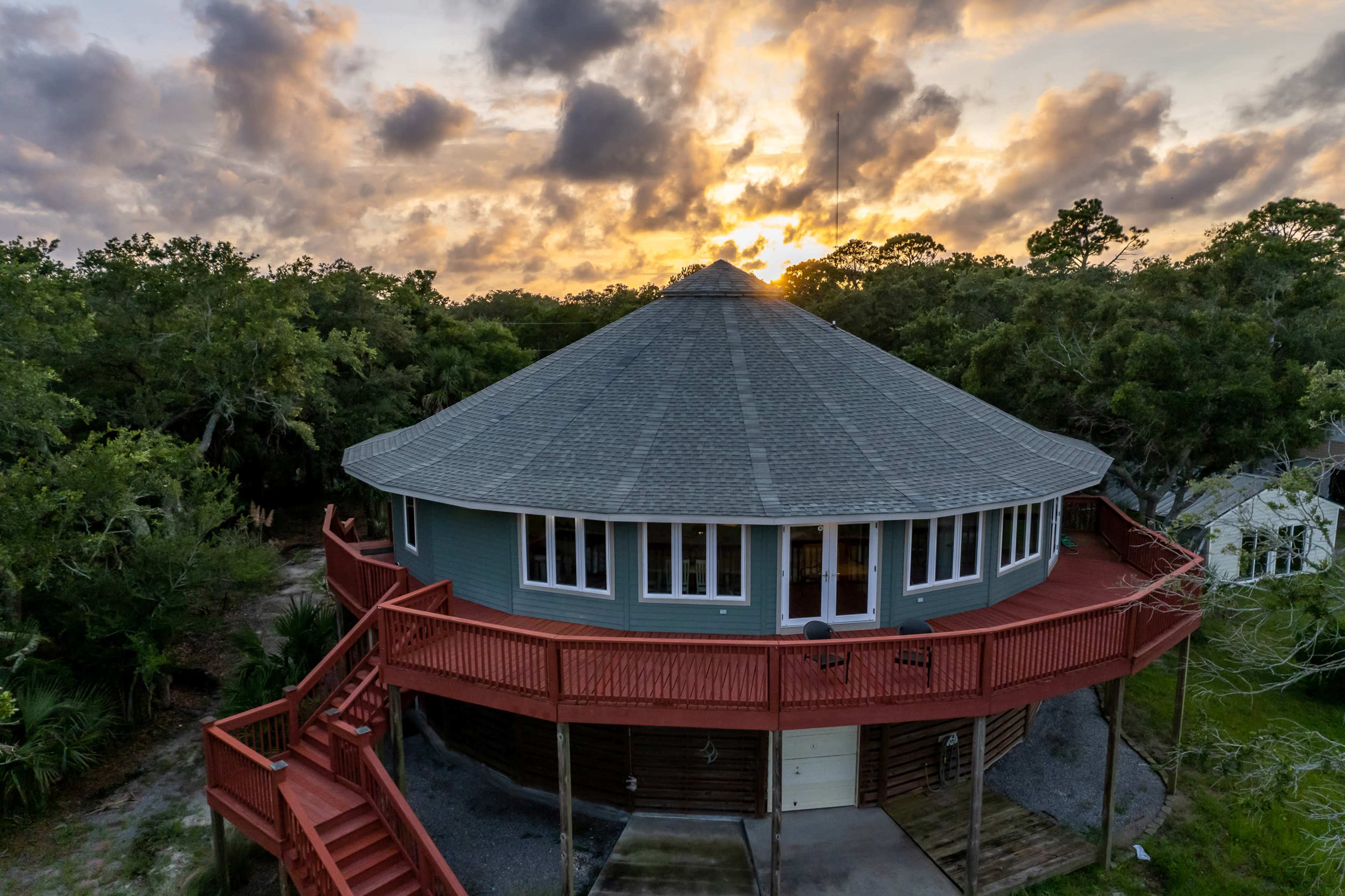 A round, elevated house with a gray roof and a red deck is surrounded by trees at sunset.