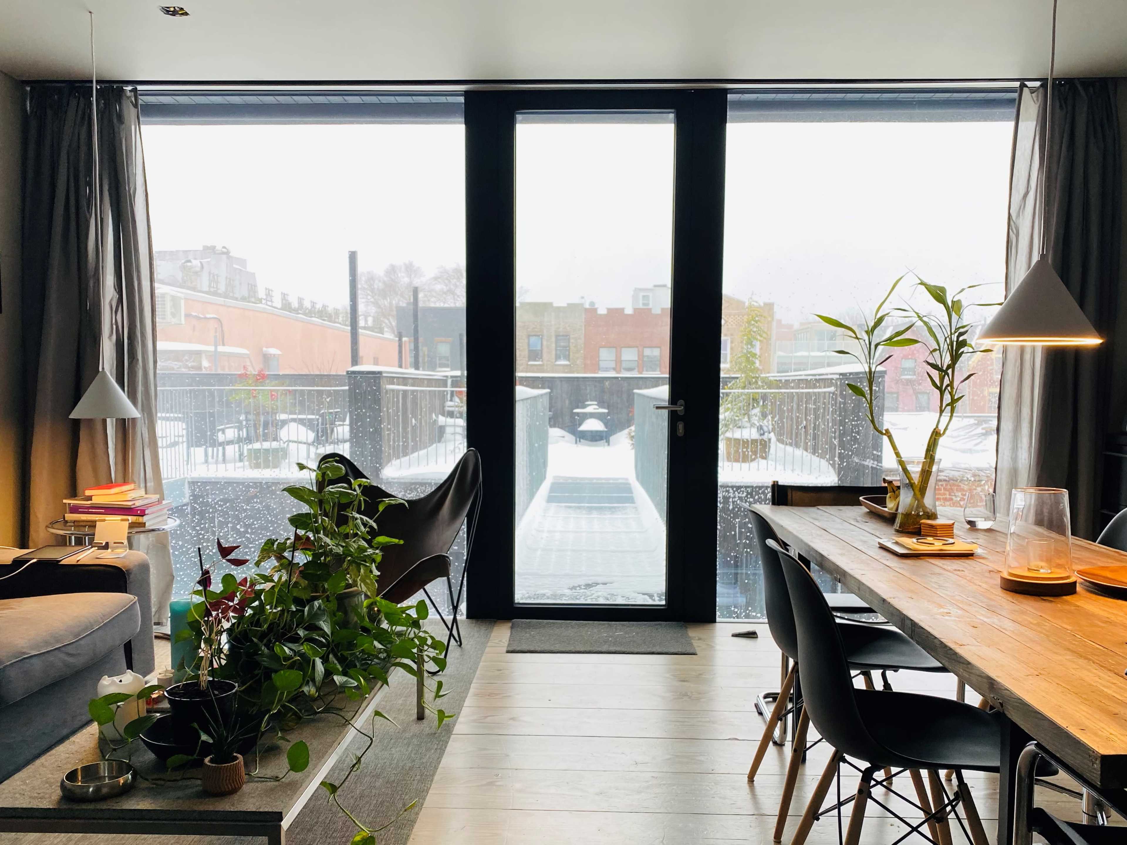 A modern living area features a view of a snowy balcony through large glass doors, with a dining table and plants positioned inside.