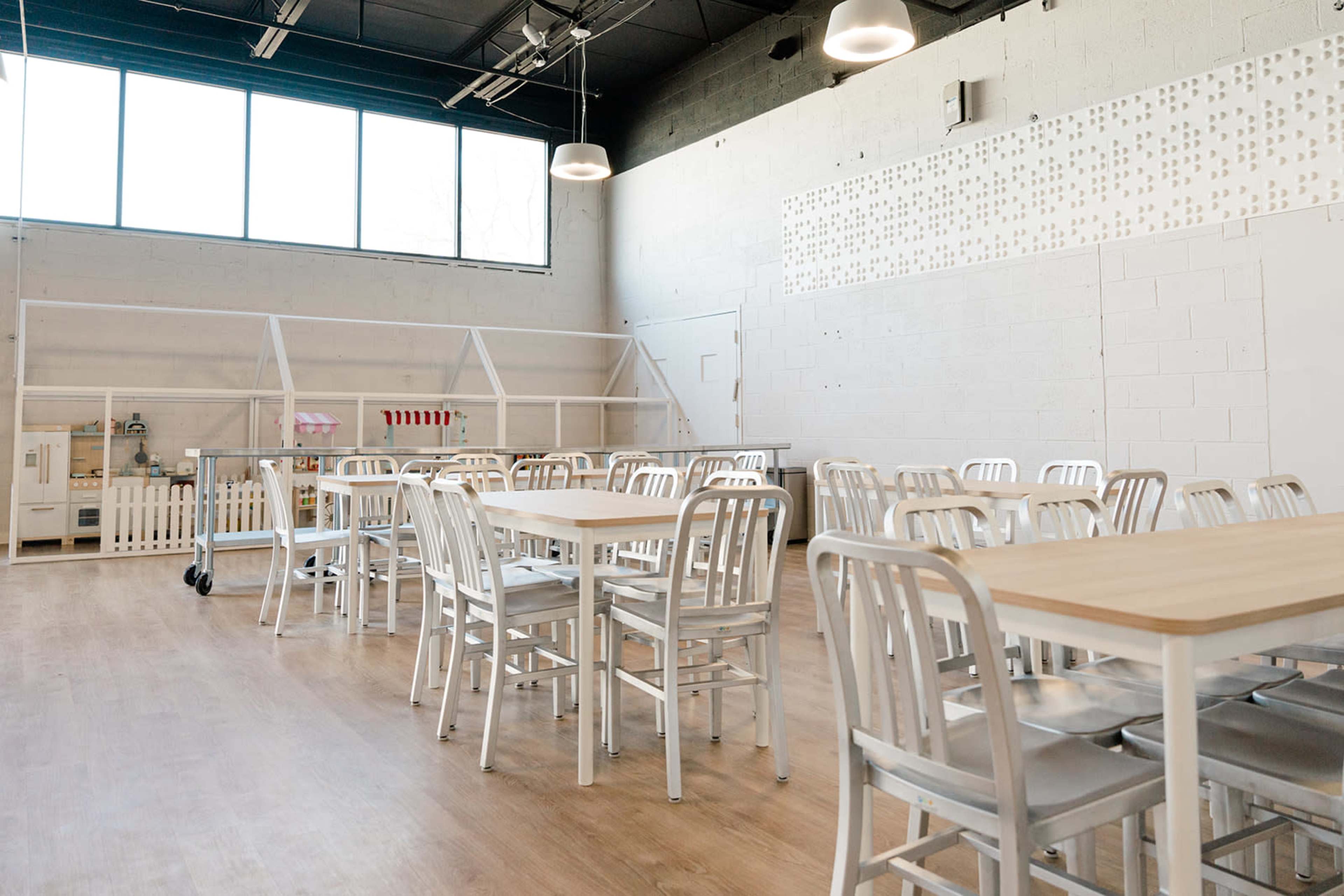 A bright, spacious room with wooden tables and metal chairs arranged in rows, along with large windows letting in natural light.