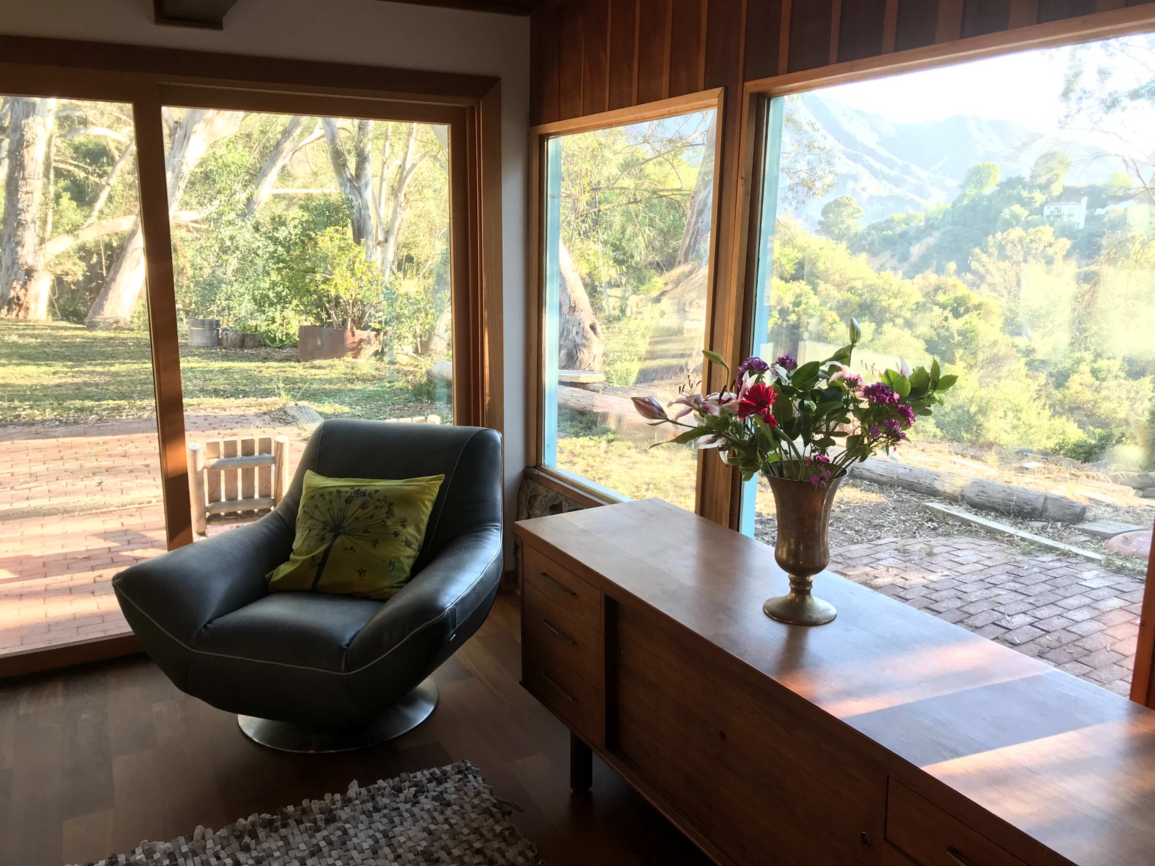A contemporary living room features a blue armchair, a wooden sideboard, and large windows overlooking a lush outdoor landscape.
