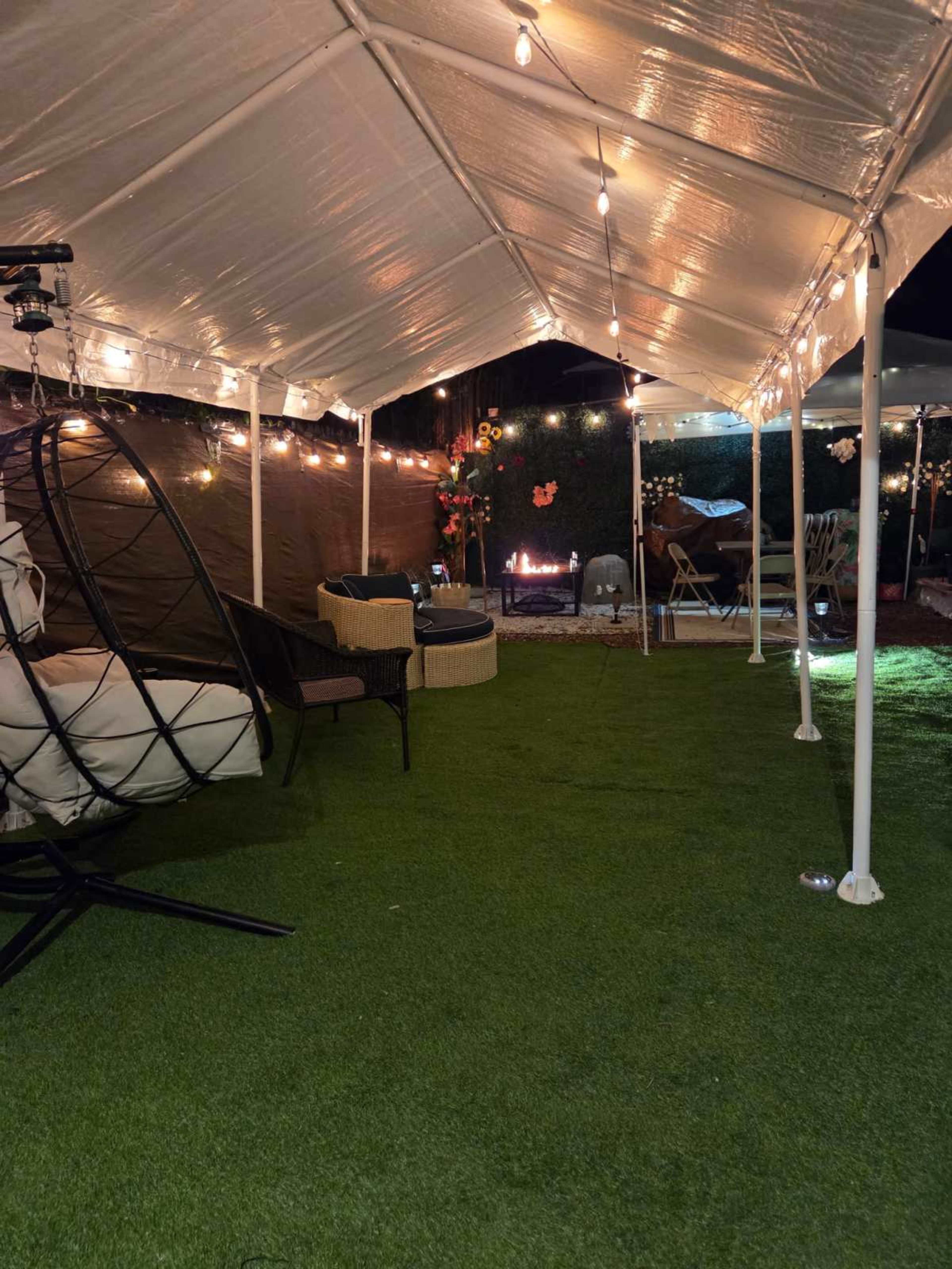 A spacious tent with string lights houses a cozy seating area featuring a hanging chair and wicker furniture, alongside a fire pit on a grassy surface.